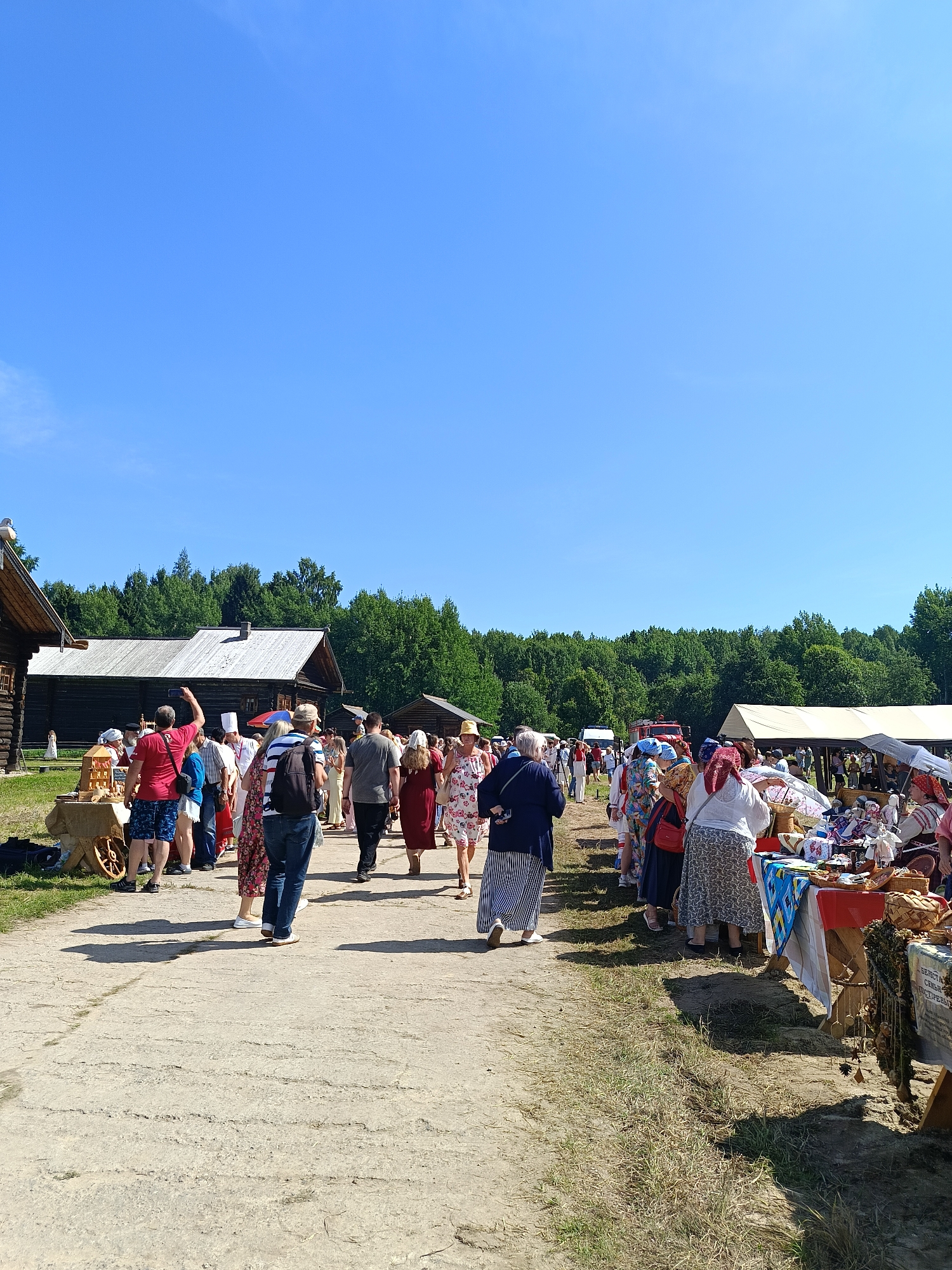 The image depicts an outdoor market or festival scene in Semenkovo, Russia, characterized by a bright, clear blue sky and a backdrop of lush green trees. A dirt path runs through the center, flanked by wooden structures on the left and market stalls on the right. A crowd of people, dressed in a variety of casual and traditional attire, are walking along the path and browsing the stalls. On the left, traditional wooden buildings are visible, hinting at a historical or cultural site. The market stalls on the right are laden with various goods, including textiles, crafts, and possibly food items, suggesting a lively commercial atmosphere. The overall scene conveys a sense of community gathering and cultural celebration.