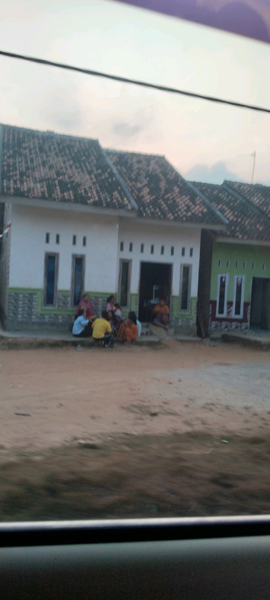 The image captures a row of houses in Tanjung Ratu Ilir, Indonesia. The houses have tiled roofs, white and green facades, and visible windows. A group of approximately six to seven people are seated in front of one of the houses, appearing to be interacting. The ground in front of the houses is unpaved and sandy. The overall scene suggests a residential area in a rural or semi-rural setting.