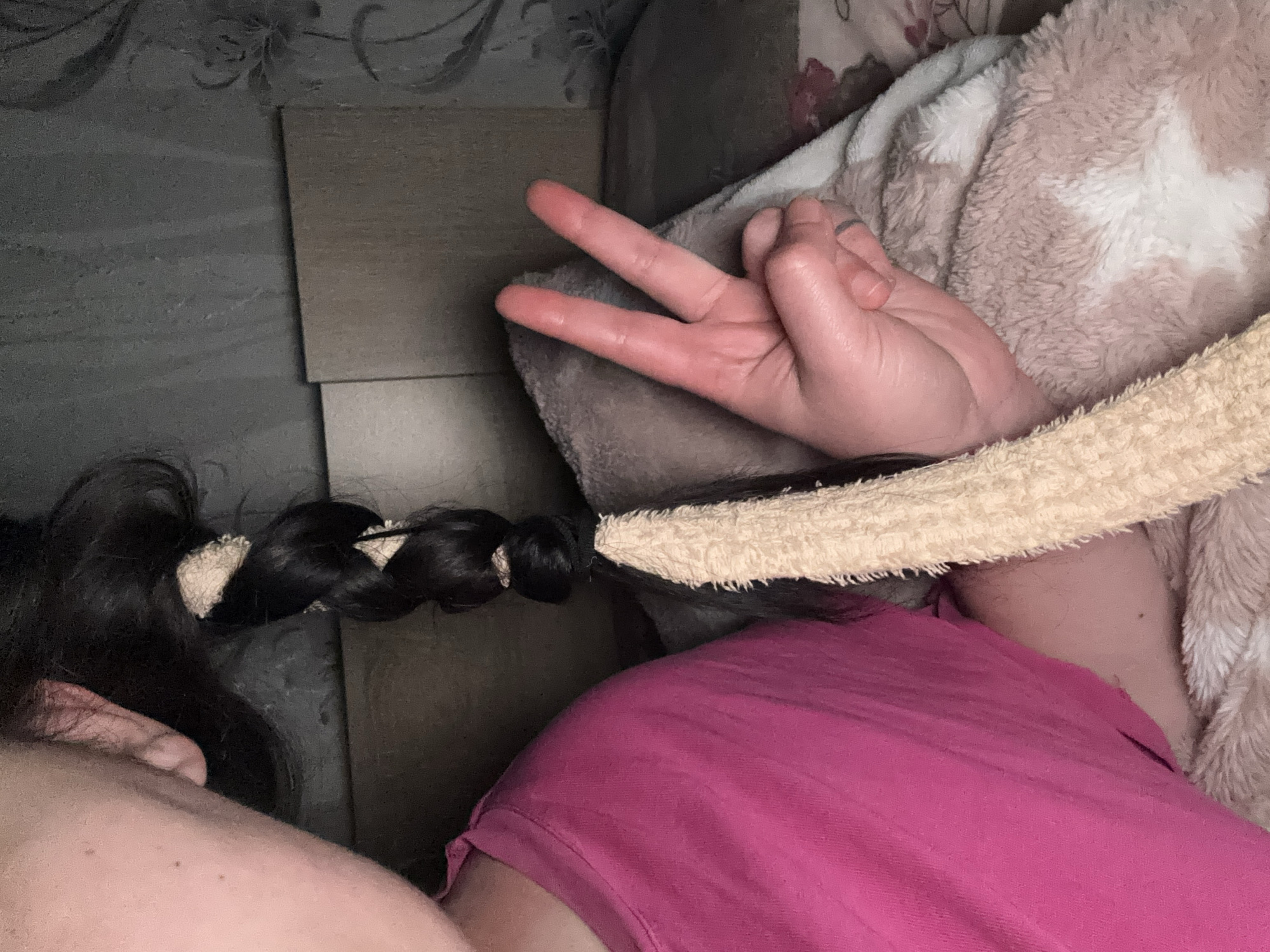 An overhead close-up shows a person lying on a surface with a pink shirt, a brown and white blanket, and a light-colored fluffy object around their neck or head. The person's dark hair is in a braid, with light-colored ties. The person's hand is making a peace sign. The background includes a wall with textured wallpaper and a tiled section. The city of Dobrovnitsa, Bulgaria is not discernible in this image.
