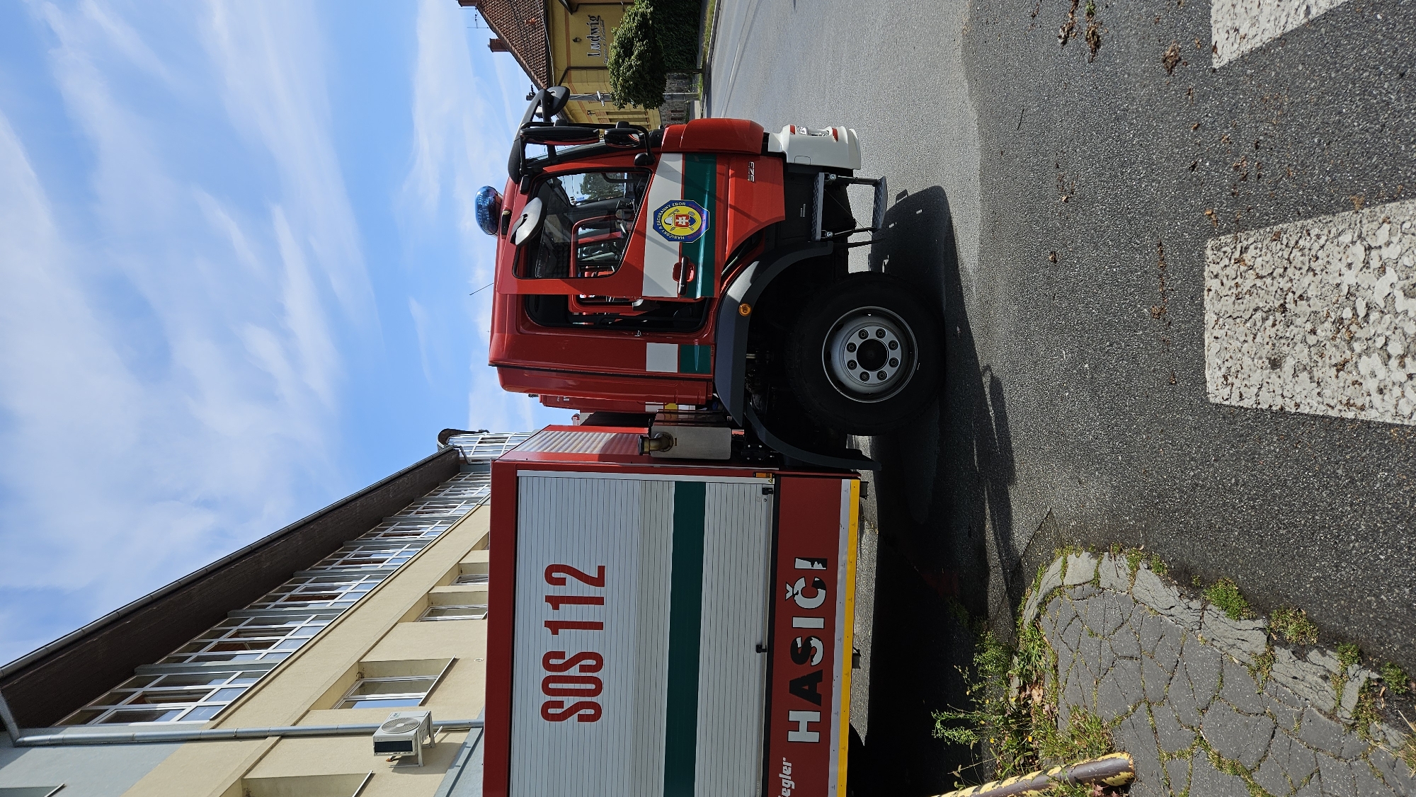 The image shows a red fire truck parked on a street in Prešov, Slovakia. The fire truck has "SOS 112" and "HASIČI" written on its side, indicating it is a fire emergency vehicle. The truck is parked in front of a building that appears to be a school or public institution, with windows running along its facade. The street has a pedestrian crossing, and there are some trees and buildings visible in the background. The sky is blue with some clouds. The scene appears calm and uneventful, with no people visible in the immediate vicinity of the truck.