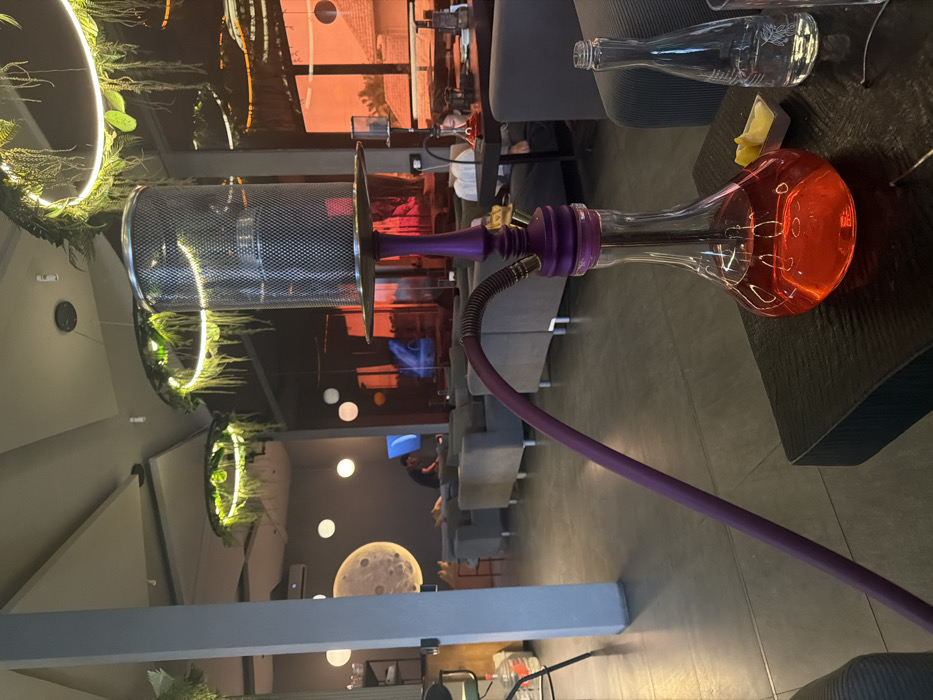 This image displays the interior of a lounge or shisha cafe, located in Tibubeneng, Indonesia, captured during evening hours. In the foreground, a shisha pipe with a purple stem, a clear glass base filled with a red liquid, and a metal mesh cover is positioned on a dark, textured low table. A purple hose extends from the shisha to the left. Adjacent to the shisha on the table are a small white dish containing two lemon wedges and an empty clear glass bottle.
The midground features multiple grey upholstered lounge sofas. A male individual in a dark shirt is seated on a sofa further back, facing a wall-mounted screen displaying a blue illuminated image. To the right, a female with dark hair is seated at a table, looking down at an object.
The environment includes a grey floor, dark grey walls, and a ceiling fitted with several circular light fixtures incorporating green foliage. A large, illuminated circular moon graphic is present on a background wall, alongside smaller spherical wall lights. Large windows on the right display an exterior view tinted orange, reflecting ambiguous outdoor structures. A white vertical pillar is visible on the left, and an air conditioning unit is mounted high on a distant wall.