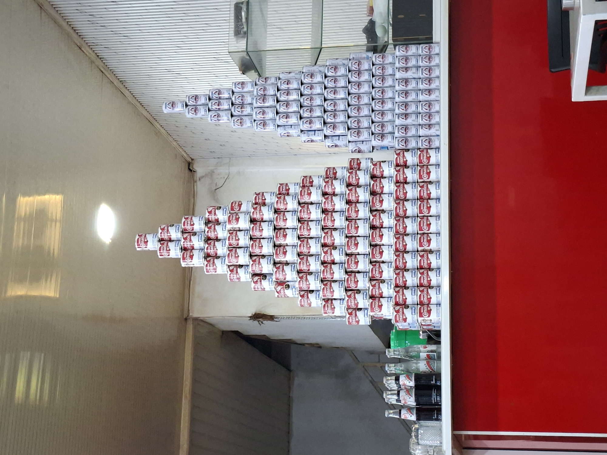 This image depicts an indoor commercial setting, likely a small shop or cafe, in Sigli, Indonesia. The central focus is a white counter with a bright red front panel, upon which two large pyramids of canned goods are meticulously stacked.
On the left side of the counter, a pyramid constructed from white cans featuring red "Carnation" branding is prominently displayed. To its right, a second pyramid, slightly taller and narrower, is formed by white cans with blue-grey accents and a distinctive crest logo.
On the left portion of the white countertop, several glass bottles, some containing dark liquid and others clear, are arranged alongside empty drinking glasses and a silver-toned metal dispenser. The background on the left consists of a plain, light-colored wall, possibly concrete, with a darker opening or stairwell receding into the distance. The background on the right features a light-colored wall with vertical corrugated or grooved paneling. A multi-tiered glass display cabinet with a dark object and a light-colored round object on its shelves is visible on the far right. The light-colored ceiling contains two recessed lights, casting reflections. No people are visible in the scene.