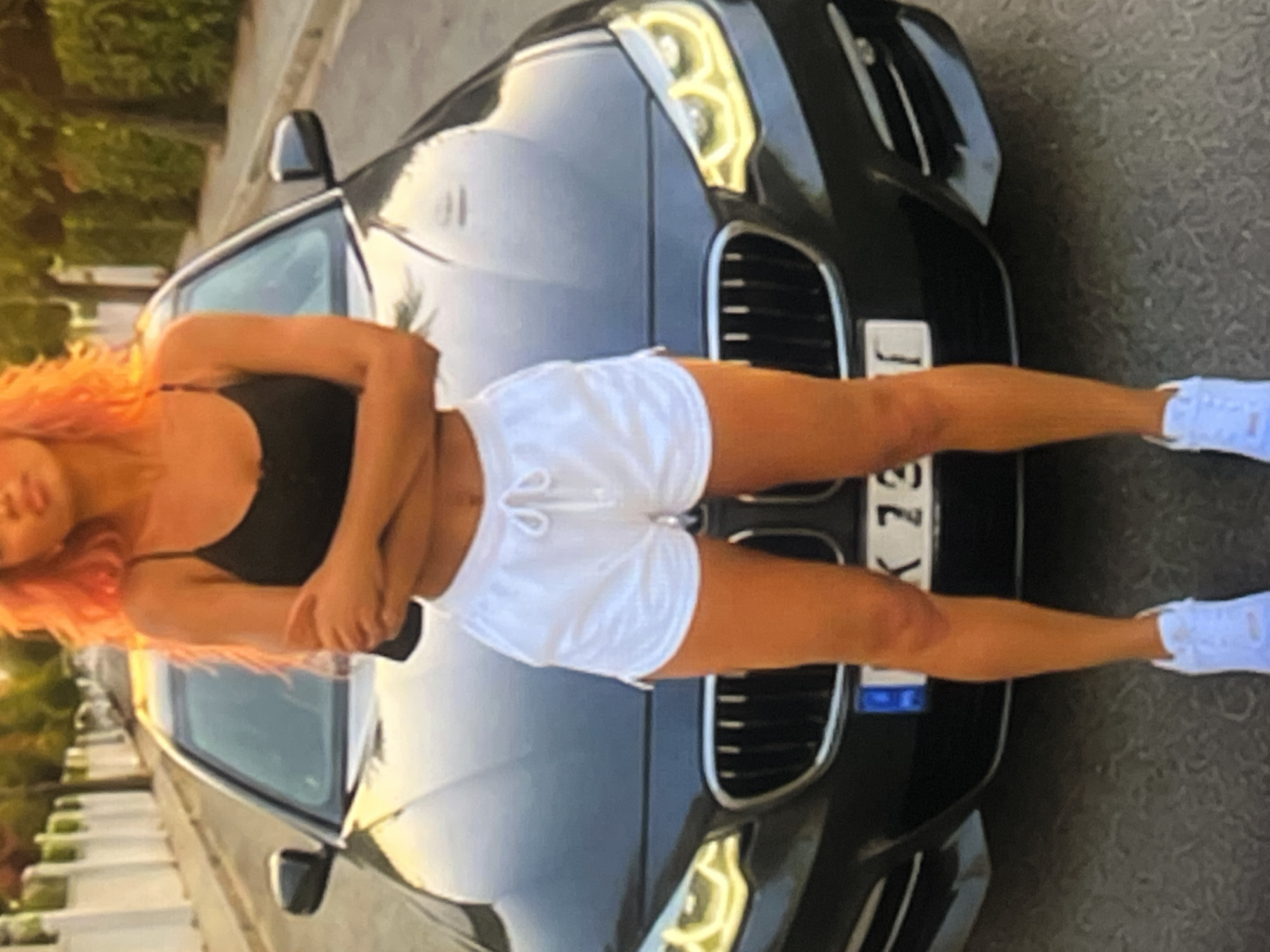 A woman with bright orange hair stands in front of a dark-colored sedan, which is parked on a street. The woman is wearing a black tank top and white shorts. She has her arms crossed and is looking down. The car's headlights are on, and the license plate is partially visible, showing "K 73". The street is lined with trees and a white fence. The lighting suggests it is either sunrise or sunset. The scene appears to be in a residential area of a city.