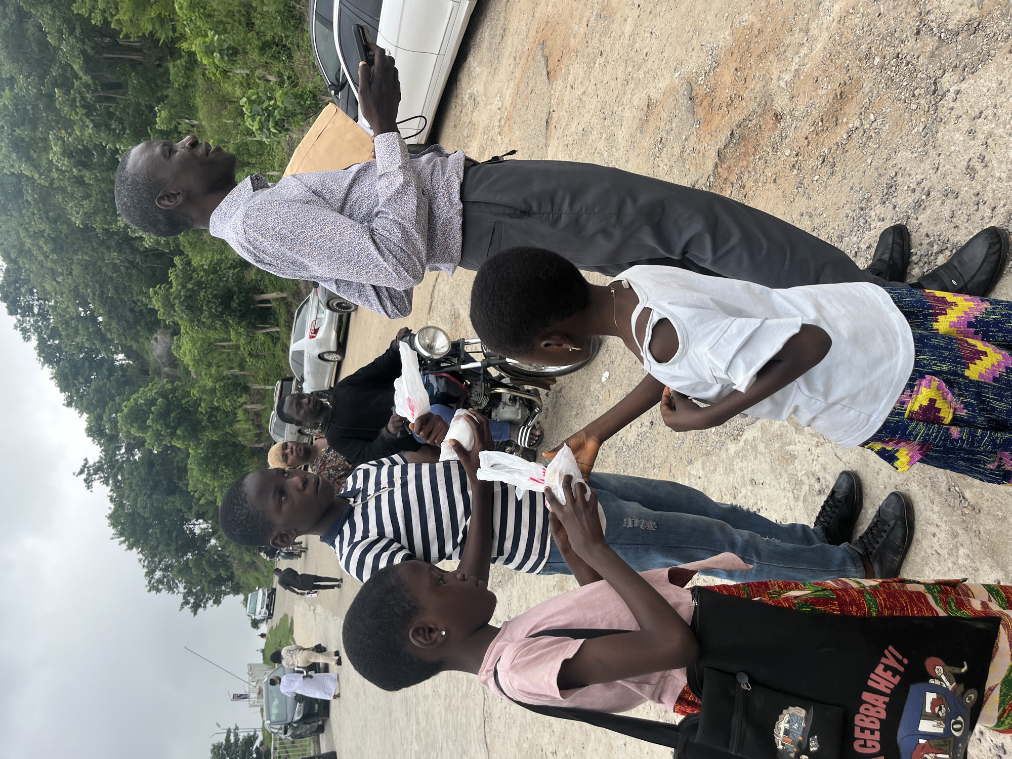 In Abuja, Nigeria, a group of people are gathered on a dusty, uneven road. Two young girls in the foreground are interacting with a boy who is holding white plastic bags, possibly food items. The girl on the left wears a pink t-shirt and a patterned skirt, carrying a black tote bag with graphics and text. The girl on the right wears a white top with ruffled sleeves and a colorful patterned skirt. The boy between them wears a striped t-shirt and blue jeans. Behind them, a man in a patterned shirt and grey trousers holds a phone and a document. Other individuals and vehicles, including a motorcycle, are visible in the background, suggesting a public area or roadside gathering. The environment is characterized by trees and vegetation on a hill in the distance under an overcast sky.