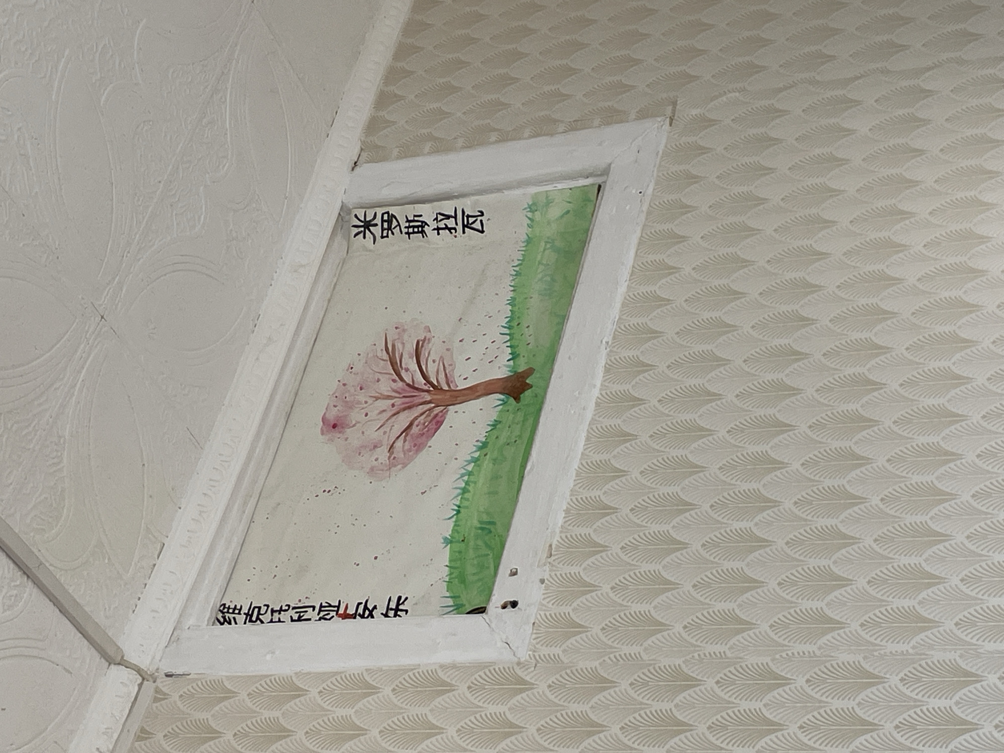 The image shows a painting affixed to the wall/ceiling. The painting is framed by a white rectangular border and features a tree with pink blossoms. Green grass is depicted along one side of the tree. Inscribed in black characters along the top and bottom of the painting are some words in what appears to be Chinese. The surrounding wall/ceiling has two distinct patterns, one with a patterned texture and the other with a more subtle feather-like design. The location is Unknown, Unknown. No people, subjects, or actions are discernible from the image.