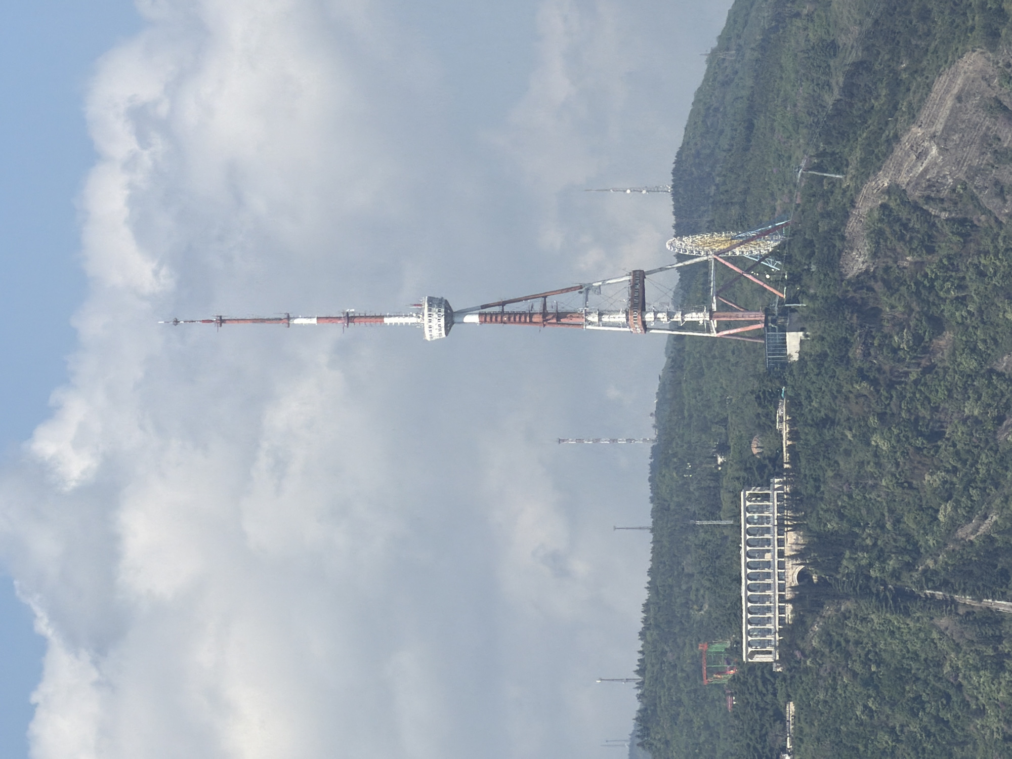The image shows the Tbilisi TV Broadcasting Tower in Tbilisi, Georgia. It is a tall, red and white structure set against a backdrop of mountains and clouds. Nearby is a Ferris wheel and other structures amidst green foliage. The image is taken from a distance, providing a broad view of the tower and its surrounding environment.