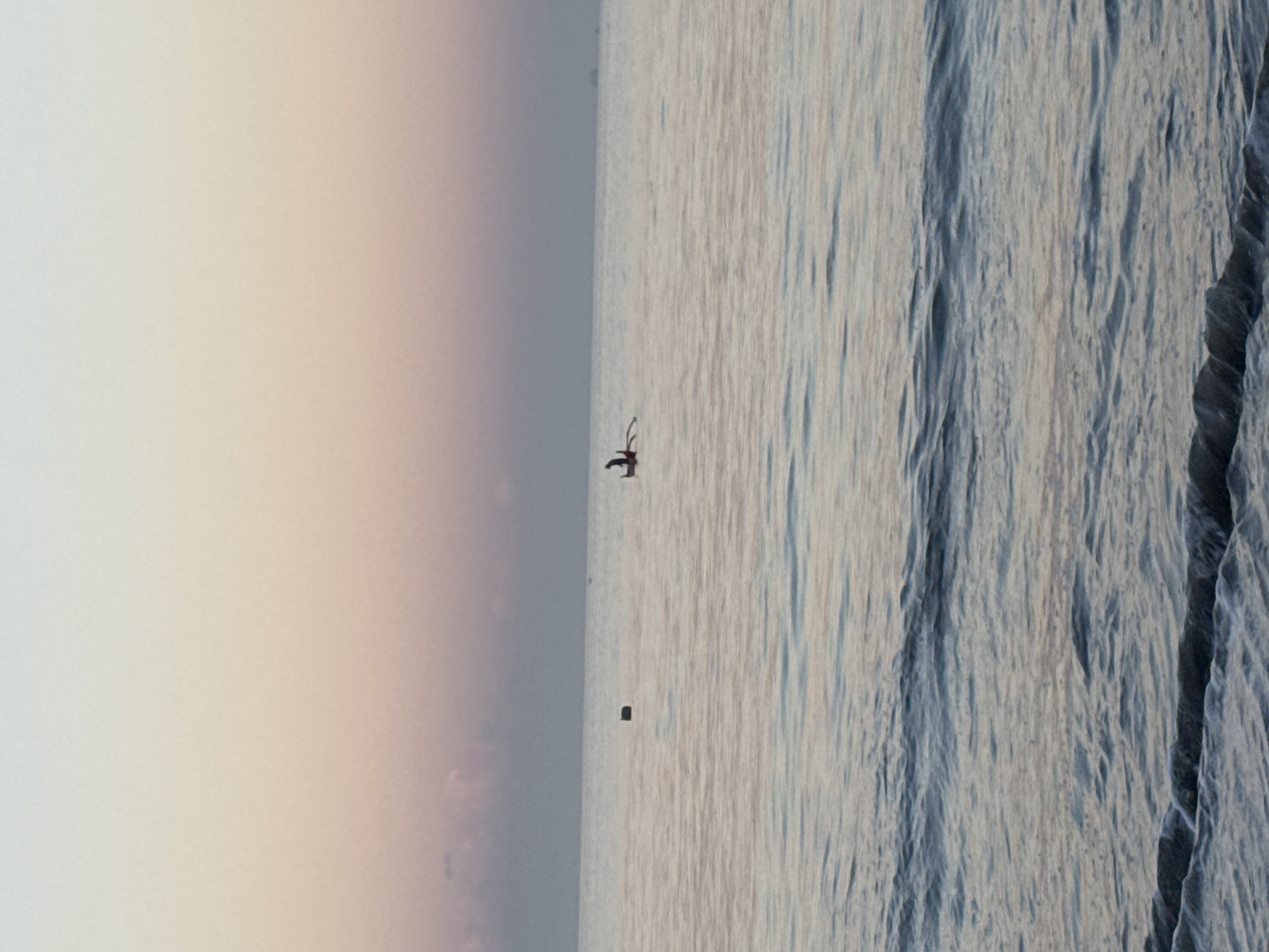 The image depicts a serene coastal scene, likely off the shores of Kuta, Indonesia, during either sunrise or sunset. The sky, occupying the left half of the frame (when correctly oriented), presents a soft gradient of colors, transitioning from a pale, almost white hue at the top to gentle tones of light lavender and faint orange-yellow towards the horizon.
The right half of the image showcases the vast expanse of the ocean. A clear, flat horizon line separates the sky from the water. In the mid-ground of the water, a single individual is visible, silhouetted against the shimmering surface. This person appears to be actively engaged in a water sport, possibly surfing or kneeboarding, as they are positioned on a board. Their dark form is clearly distinct against the brighter water, though specific details of their attire or equipment are obscured by the distance and backlighting.
Further out in the ocean, a smaller, dark, rectangular object floats. Its precise nature is indistinct, but it could be a buoy, a small vessel, or debris. The water in the foreground exhibits a textured surface with ripples and gentle waves, reflecting the soft light and creating variations in tone, predominantly in shades of greyish-blue. The overall impression is one of tranquil natural beauty and solitary activity on the open water under diffused light conditions. There is no visible text in the media file.