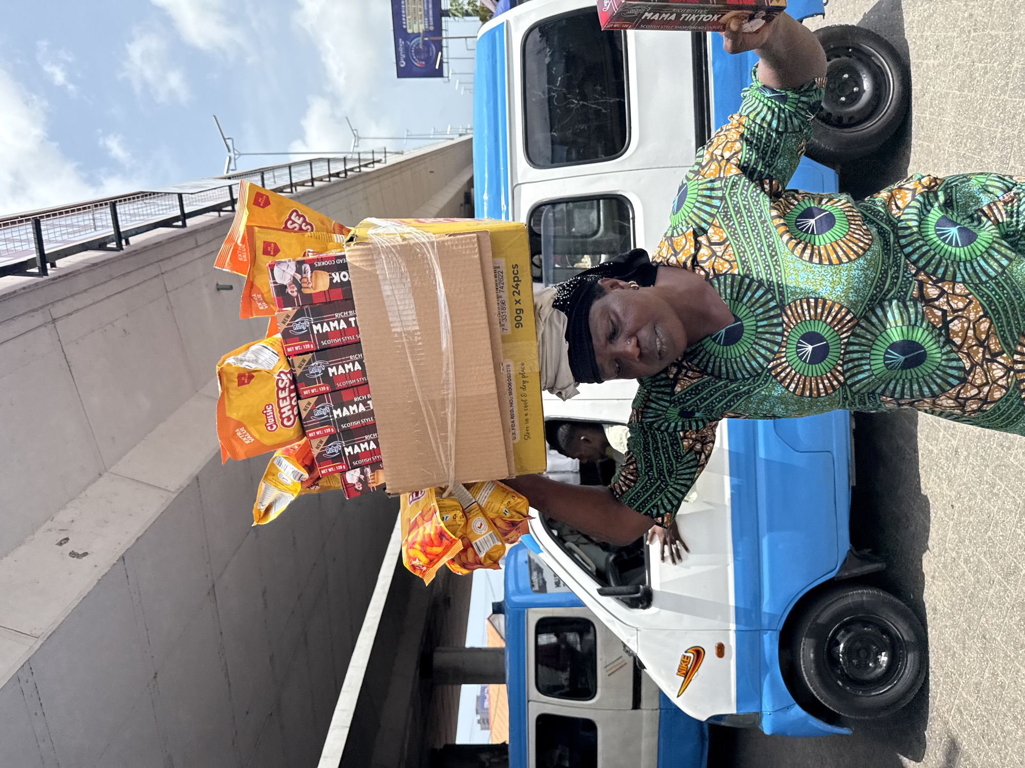 The image depicts a woman in Lagos, Nigeria, selling goods. She is wearing a green and brown patterned dress and carrying a stack of items on her head, including boxes of "Ruby's Mama TikTok" Scottish style shortbread cookies and bags of cheese-flavored snacks. The vendor is positioned next to a blue and white commercial mini-bus, with the "Nike" logo displayed on its front side. The bus is located on a paved surface, possibly a street or roadside. In the background, a concrete overpass and a billboard can be seen. The scene is well-lit, indicating it is daytime. Another individual is partially visible inside the bus.