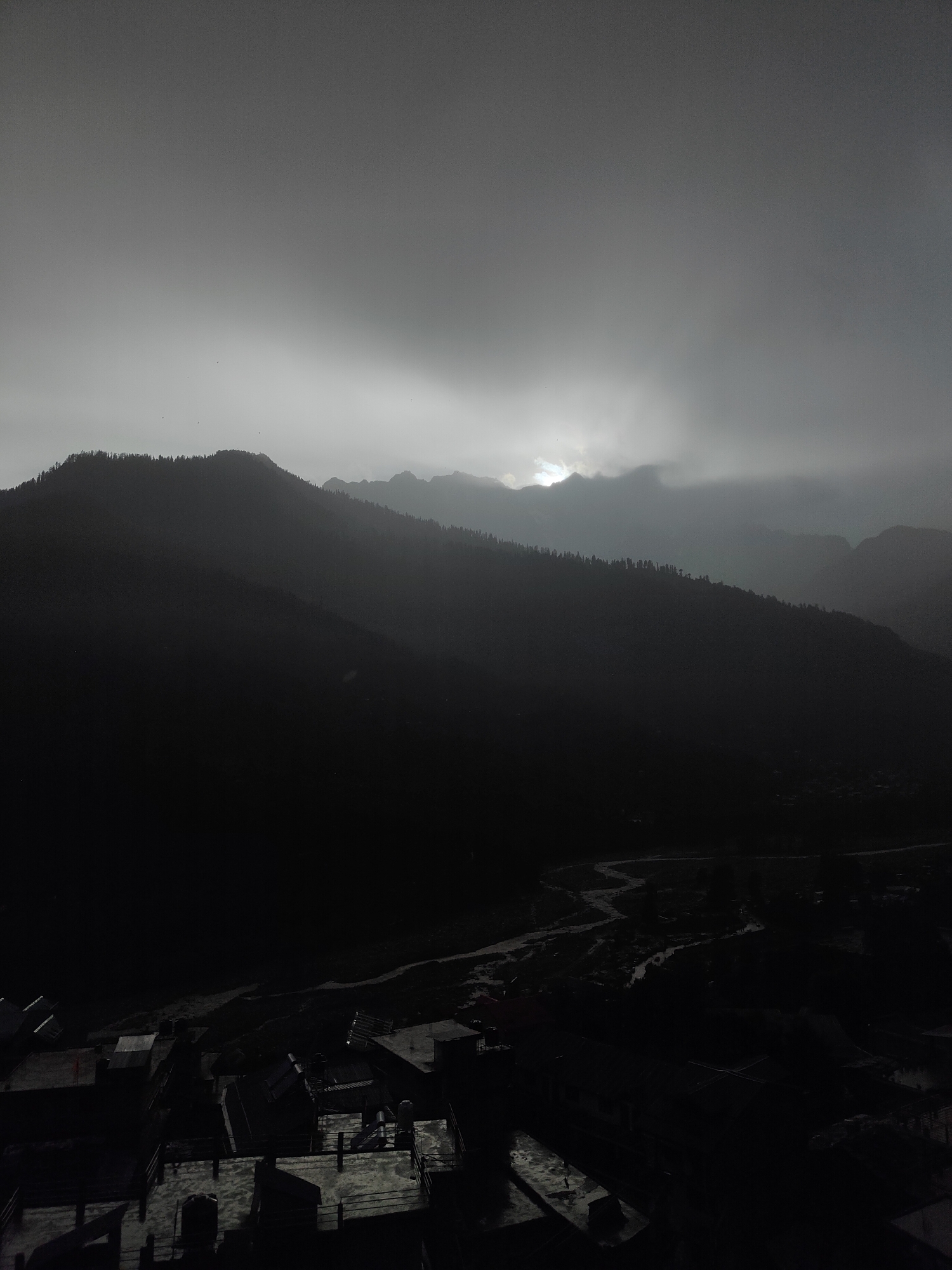 The image captures a mountainous landscape with a village nestled in the valley, appearing to be at dusk or during a heavy overcast. The sun is partially visible through thick clouds, casting a diffused light over the scene. In the foreground, there are rooftops of buildings, suggesting the perspective is from an elevated position within the village. A winding river or stream is visible in the middle ground, flowing through the valley. The background is dominated by dark, silhouetted mountains covered in trees. The overall atmosphere is somber and muted due to the low light conditions and the monochromatic palette of the image.