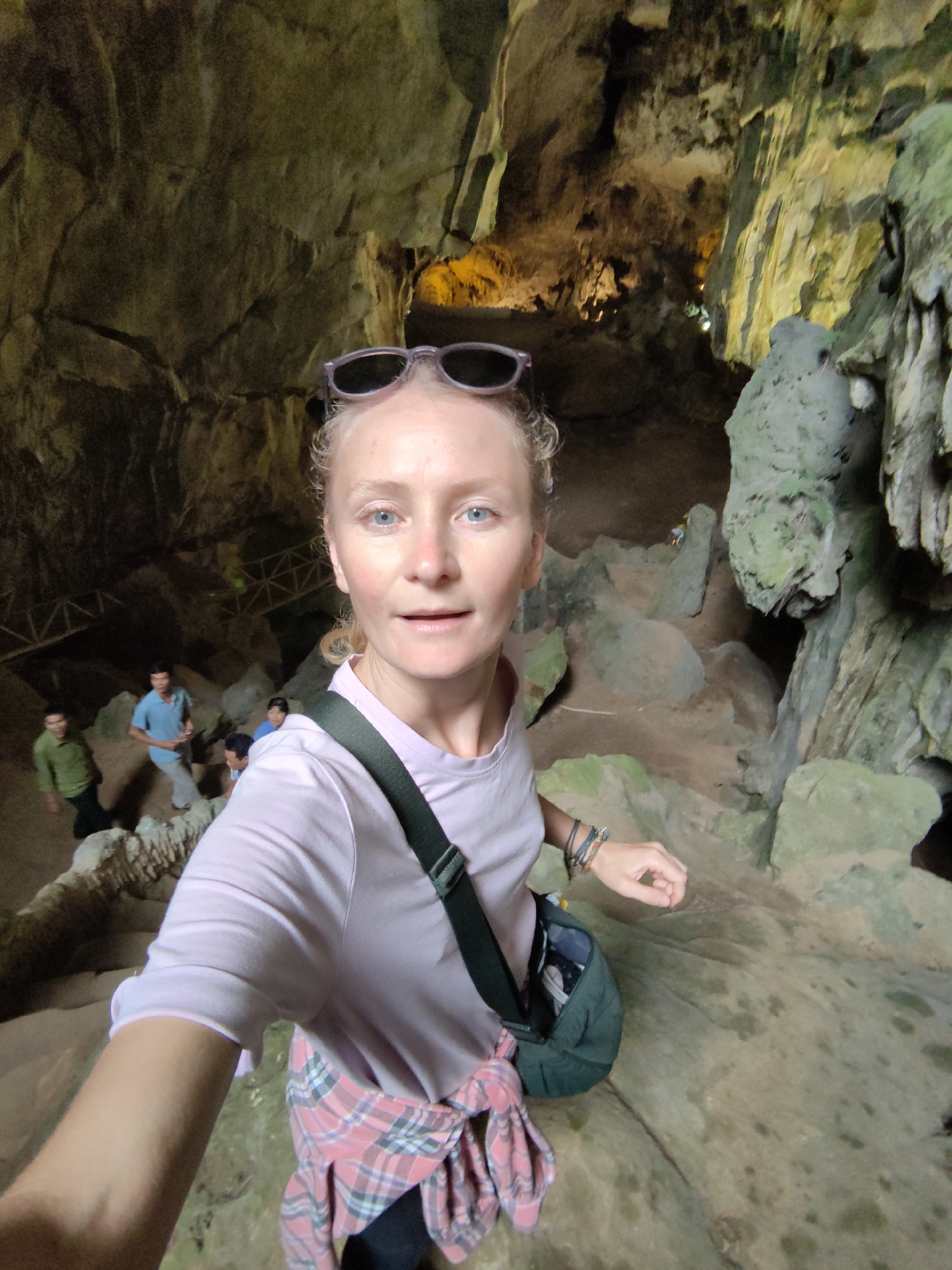 A fair-skinned woman with blonde hair pulled back, blue eyes, and wearing sunglasses on her head is taking a selfie inside a cave. She is wearing a light pink T-shirt, and a pink and gray plaid shirt is tied around her waist. A green bag with a black strap is slung across her body.
In the background, other people are visible descending a set of stone stairs with a metal railing. The cave walls are rough, textured, and appear to be covered in moss or algae. The lighting is dim with some areas illuminated by artificial light. The scene is likely inside Tam Thanh Cave in Lạng Sơn, Vietnam, known for its limestone formations and historical significance.
