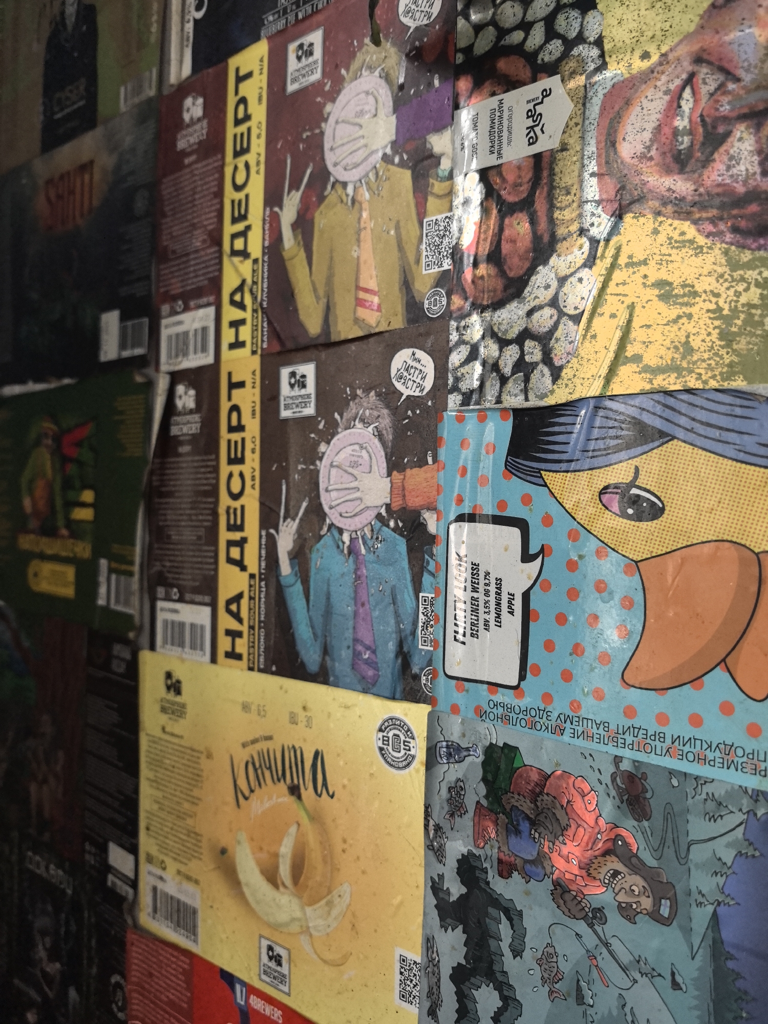 The image displays a close-up, angled view of various commercial product packaging, predominantly craft beer labels and boxes, densely arranged on a vertical surface in what is stated to be Noviy, Russia. The packaging features a range of graphic designs, including cartoon characters, stylized figures, and food illustrations. Russian text is prominent on most visible items, alongside some English descriptions.
Specific details include:
* Multiple brown and yellow packages labeled "НА ДЕСЕРТ" (FOR DESSERT), indicating "PASTRY SOUR ALE" with ABV 6.0%. These depict stylized figures in suits with plates obscuring their faces, making a "rock on" hand gesture. One variant mentions "BANANA STRAWBERRY VANILLA" and another "MILK CINNAMON COOKIES."
* A blue package with red polka dots and a partial cartoon face, labeled "ФЛИТТИЙ ЛУК" (FLIRTY LOOK), identifies as a "BERLINER WEISSE" (ABV 3.5%) with "LEMONGRASS" and "APPLE" flavors.
* A yellow package displays a partially peeled banana and the Russian word "Кончита" (Konchita), with an ABV of 6.5% and IBU 30.
* A dark grey/black package with a patterned design holds a white label with "ОГОРОДИЩЬ: МАРИНОВАННЫЕ ПОМИДОРКИ" (MARINATED TOMATOES).
* A package on the lower right features a colorful illustration of a robust, bearded man in an orange jacket, fishing in a forest setting with trees and fish. Text at the top includes a Russian warning about alcohol consumption.
* Visible brand names include "Atmosphere Brewery" and "4Brewers."
* The packaging appears slightly creased or worn in places, indicative of display or storage. No individuals are directly visible in the scene, only depicted characters on the packaging.