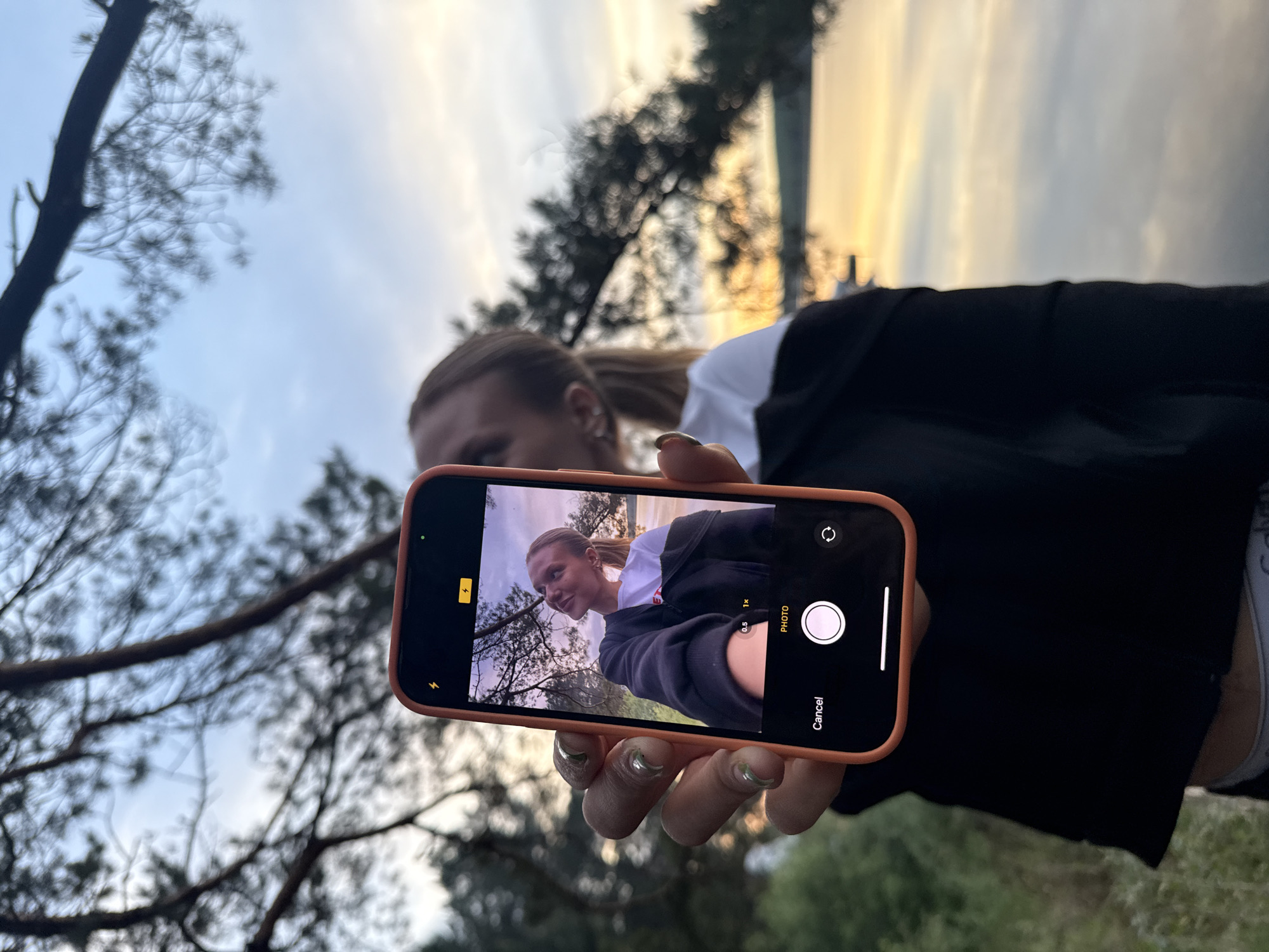 A photograph depicts a person holding a smartphone displaying a photo of themselves in a similar setting, likely taken moments prior. The person holding the phone is partially visible in the background, appearing to be taking a "photo in photo" shot. The location appears to be an outdoor, wooded area with trees and foliage visible in the background, and a muted sky with golden light. The exact city is not discernible from the image.