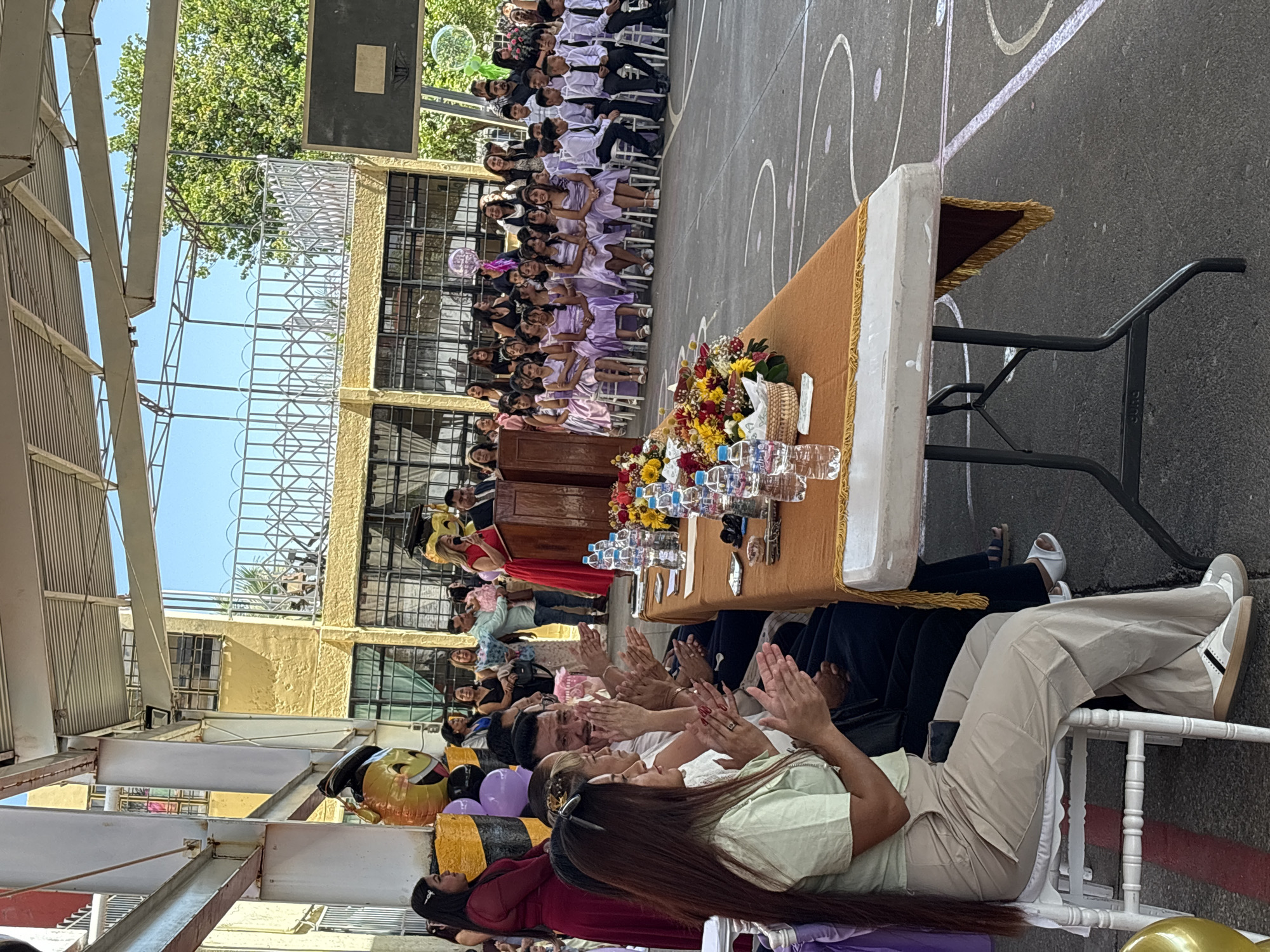 The image depicts an outdoor graduation ceremony or school event taking place in Tejalpa, Mexico, under a covered structure. A woman in a red dress stands at a wooden podium, speaking into a microphone. In front of her, a long table is laden with floral arrangements and bottled water. Spectators, including students in uniform and adults, are seated or standing. Balloons, including one shaped like a bee, are visible. The background shows a school building and trees.