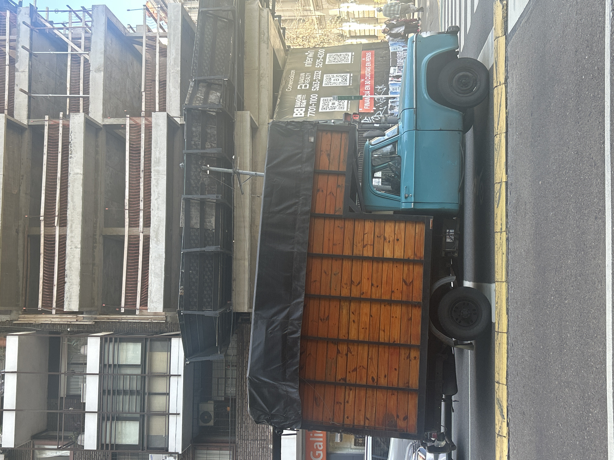 The image depicts a turquoise vintage truck with a wooden cargo bed covered by a black tarp, parked on a street in Buenos Aires, Argentina. The truck is in motion, crossing a pedestrian intersection marked by yellow and white lines. In the background, a building under construction is visible, with scaffolding and concrete structures. To the left of the truck, another building with balconies and windows is present. A large banner advertising real estate services, including QR codes and phone numbers, is attached to the construction site. Several people are walking on the sidewalk in the background. The scene appears to be in a commercial or urban area of the city.