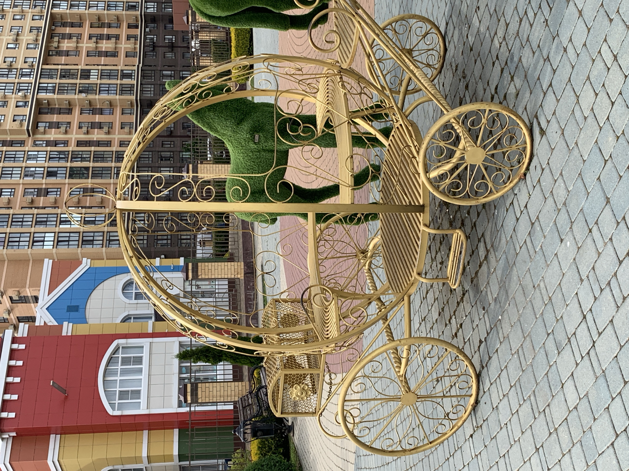 A golden ornate carriage, resembling a Cinderella carriage, is positioned on a paved surface in front of a colorful building and a tall, modern apartment building. The carriage is elaborately decorated with swirls and curves, featuring two seats and two decorative wheels. Inside the carriage, two green, topiary horses are present. The building in the background is a vibrant mix of red, yellow, and blue with arched windows and white trim. Beyond that, a multi-story brick apartment building with numerous windows is visible. The paved ground is made of gray rectangular stones.