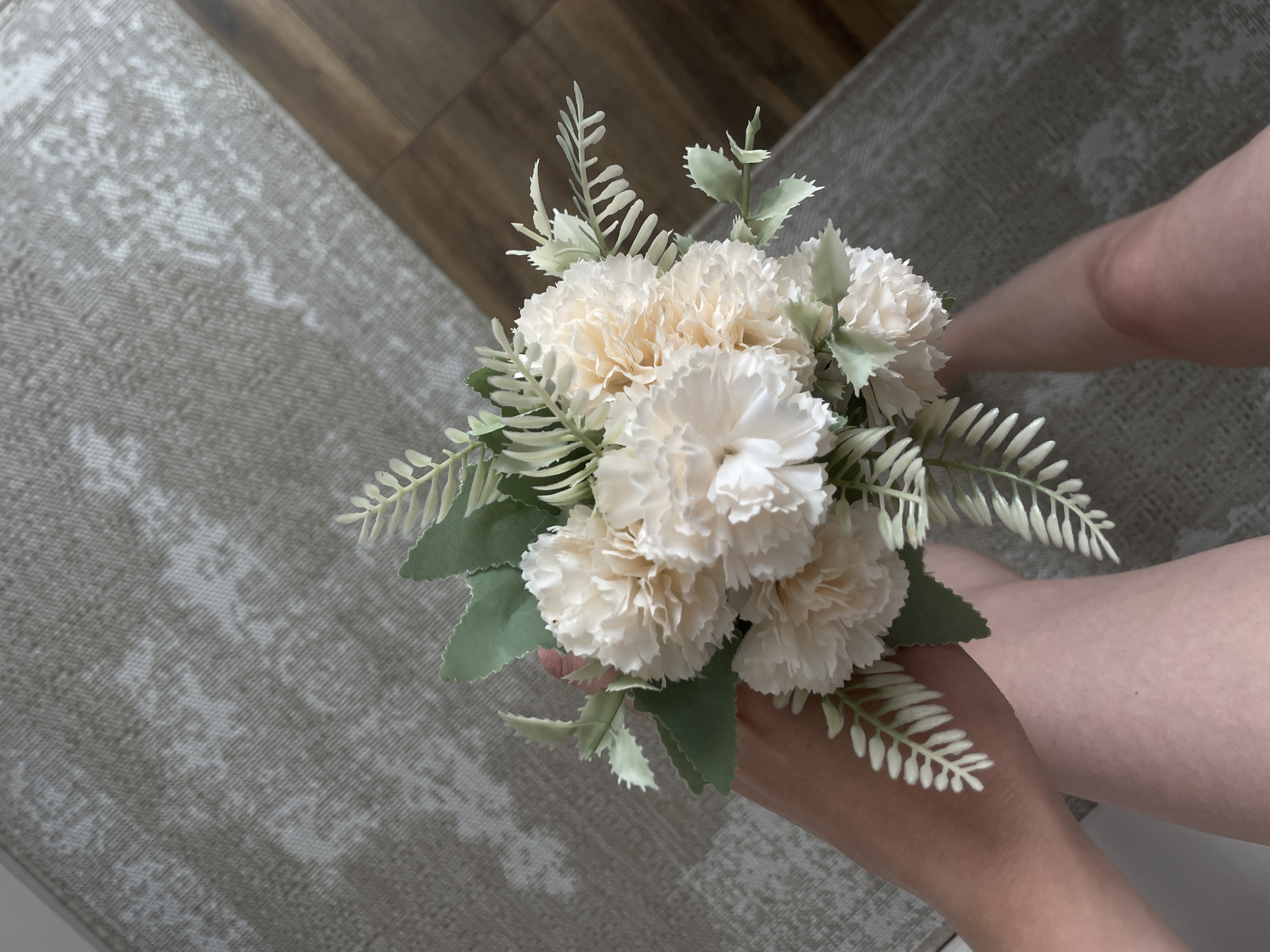 A person holds a bouquet of cream-colored flowers with interspersed greenery. The background includes a patterned rug and a wooden floor. The person's legs are visible to the right of the bouquet.