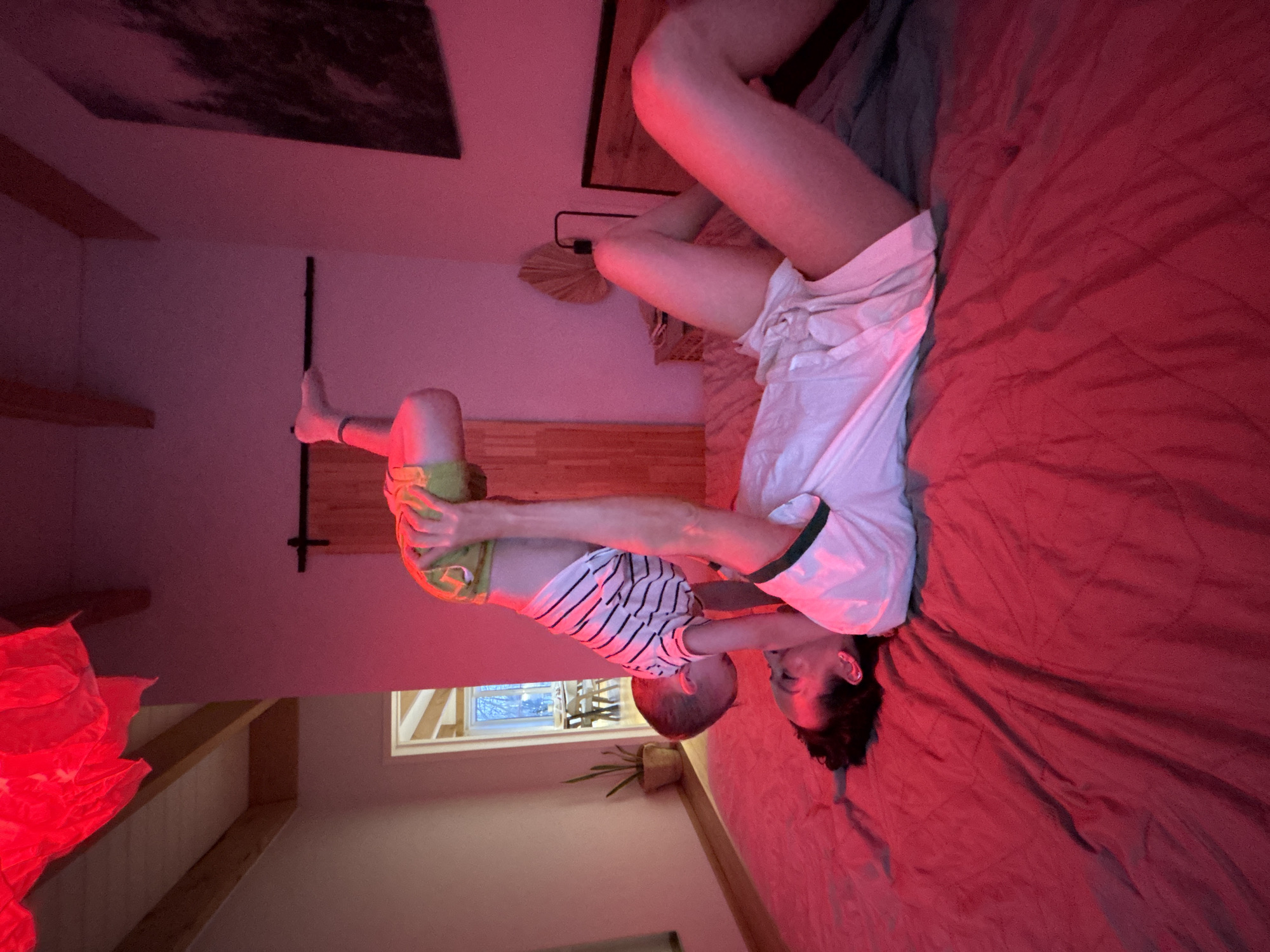 The scene depicts an adult lying on a bed covered with red sheets, interacting with a young child. The adult is positioned horizontally across the frame, wearing a white top. The child is held by the adult, wearing a striped shirt and green shorts, their feet extended upwards. The setting appears to be indoors, possibly a bedroom, with a window and a red light source illuminating the room. Other objects present include a plant, a bed, and a piece of artwork. The location is unconfirmed to be in Chaikovsky, Russia.