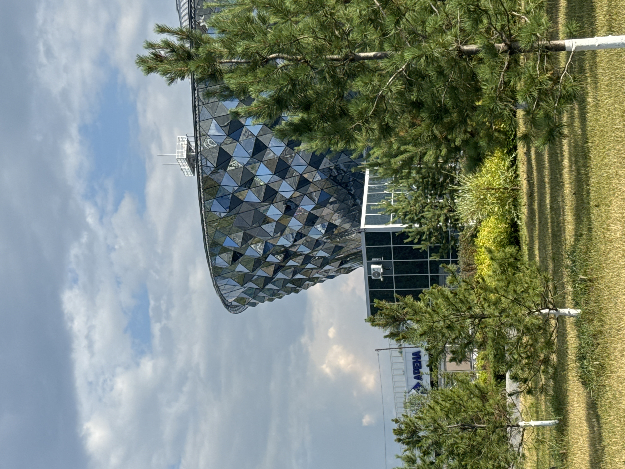 An outdoor, medium shot shows the modern, geometric facade of the "Arena" building in Novosibirsk, Russia, partially obscured by trees. The building's exterior is covered in reflective triangular panels, creating a faceted, diamond-like appearance. A blue sign reading "AREHA" is visible on the lower section of the building. In the foreground, a grassy lawn with visible shadows from sunlight is dotted with young pine trees, some of which have white-painted trunks. The sky is partly cloudy, with patches of blue sky visible through the clouds. The overall scene is one of contemporary architecture set within a landscaped, natural environment.