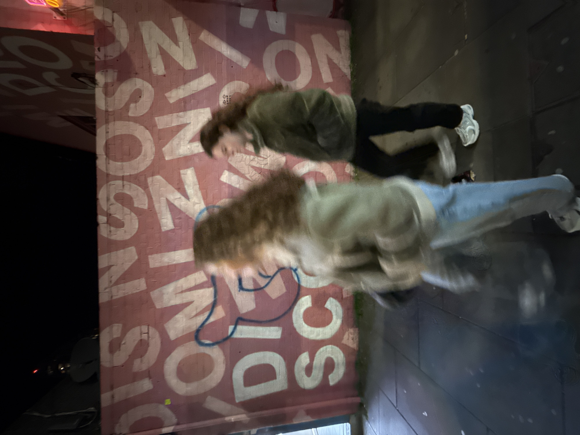 The image depicts two blurry figures walking past a wall covered in graffiti. The graffiti includes the words "SWISSCOTTAGE" in large white letters on a pink background. The figures are blurred due to motion. One figure appears to be wearing a green jacket and black pants, and the other appears to be wearing a light-colored jacket and blue jeans. The location is likely in the Swiss Cottage area of London, United Kingdom. The scene appears to be at night.