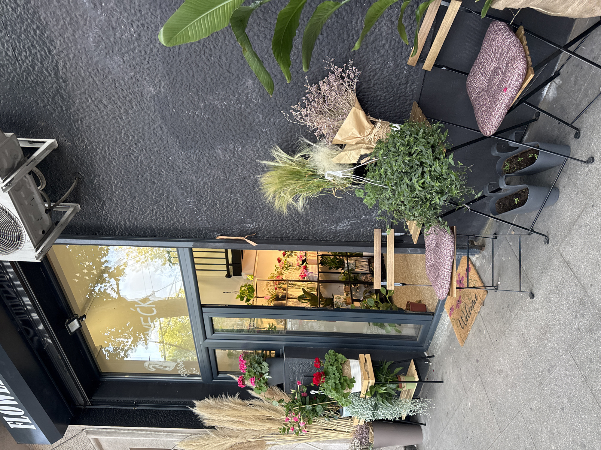 This is an eye-level, outdoor shot of a flower shop in Belgrade, Serbia. The shop has a modern aesthetic with a dark textured facade. A large glass window with a dark frame showcases the interior of the store, where an assortment of plants and flowers are visible. On the exterior, bouquets of dried flowers, potted plants, and cut flowers are arranged to attract customers. A small welcome mat lies near the shop's entrance. Outside the shop, two small chairs are placed next to each other. A leaf overhangs the top left of the shop.