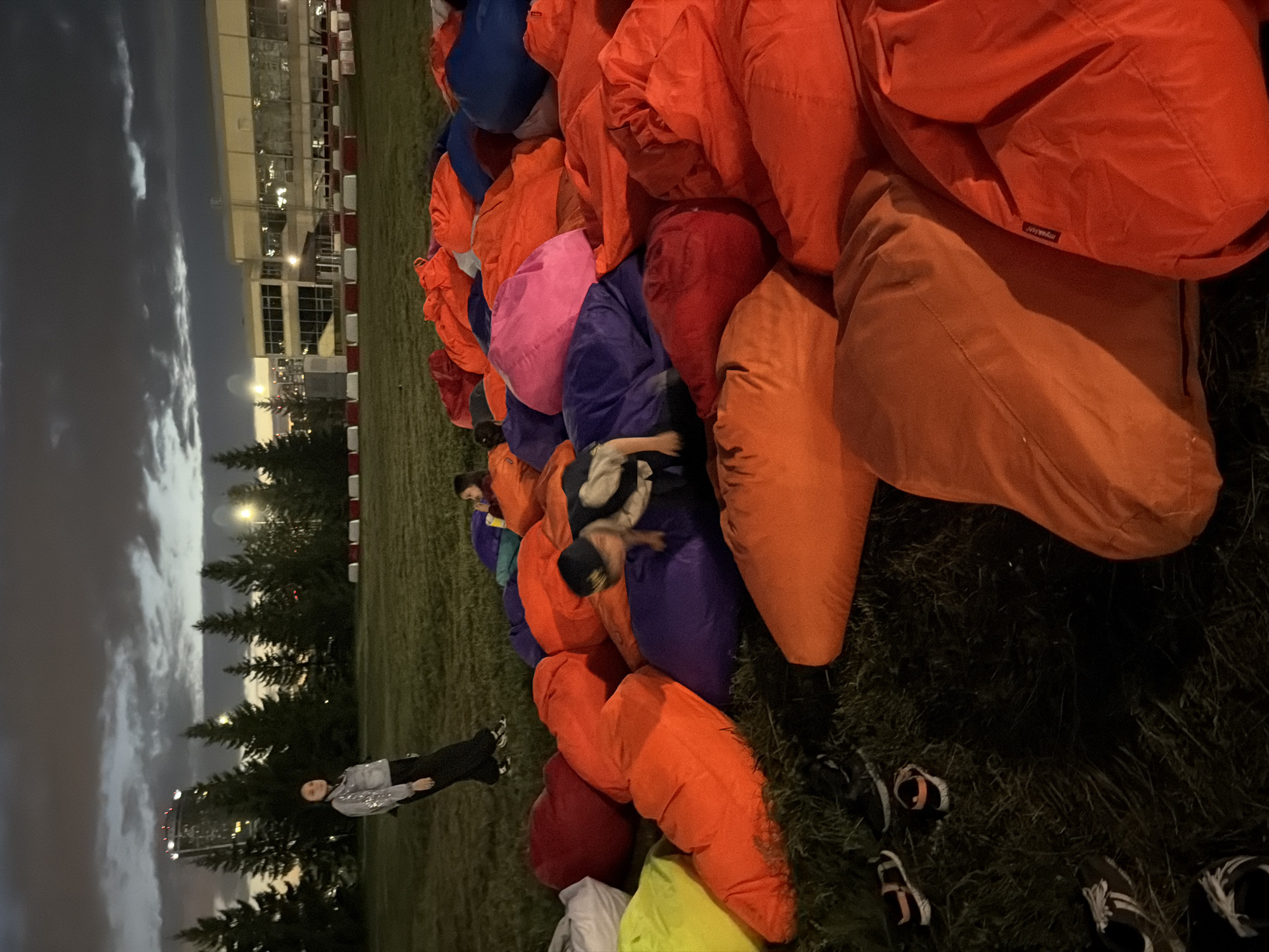 In a grassy, outdoor location at dusk, a cluster of people is gathered around a large collection of brightly colored beanbag chairs. A young boy, wearing a baseball cap, is crawling on a purple beanbag, reaching towards something out of frame. To his left, a young girl in an orange shirt holds a drink and looks towards the boy. Further in the background, a woman in a sparkly silver jacket and black pants stands on the grass, looking to her left. The background shows a line of evergreen trees, a tall building with lights on, and a building with large windows and a white and red striped barrier in front of it. The sky is cloudy and dark. The ground is covered in grass and scattered with shoes.