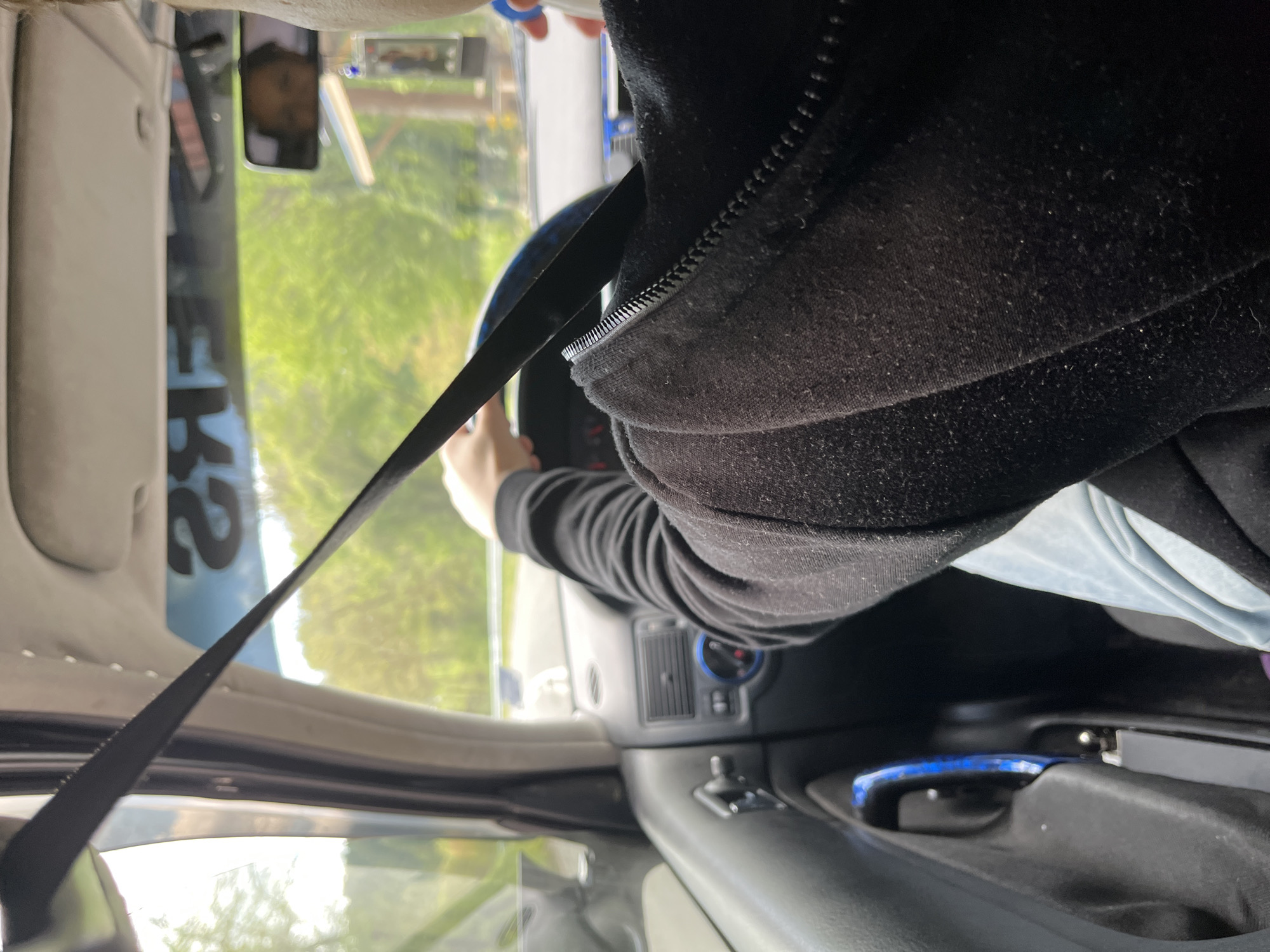 A person wearing a black hooded sweatshirt is in the driver's seat of a car, wearing a seatbelt. The person has their hands on the steering wheel. Outside the car, green trees and foliage are visible. There is a mobile phone in a holder attached to the dashboard. The location is Startsevo, Bulgaria.