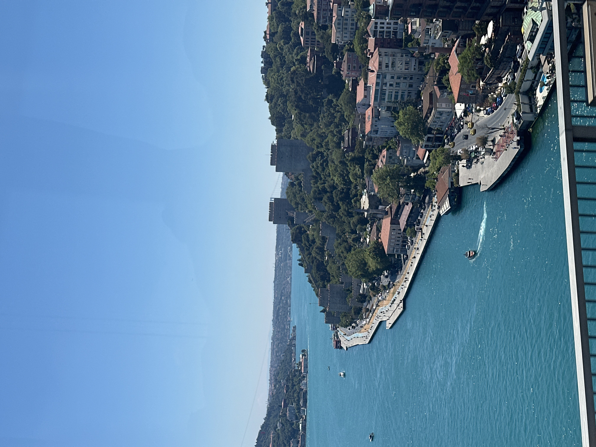 The image captures a panoramic view of the Bosphorus Strait in Sarıyer, Turkey. On the right bank, a historic stone fortress with two prominent towers stands on a verdant, tree-covered hill. Below the fortress, a coastal road is visible, lined with buildings featuring red-tiled roofs. Several vehicles, including yellow taxis, are on the road. A promenade runs along the water's edge, with pedestrians visible. Boats dot the bright blue water of the strait. In the distance, the cityscape continues, with more buildings and hills under a clear blue sky. The foreground shows a metal railing, suggesting the photo was taken from an elevated vantage point, possibly a balcony or bridge.