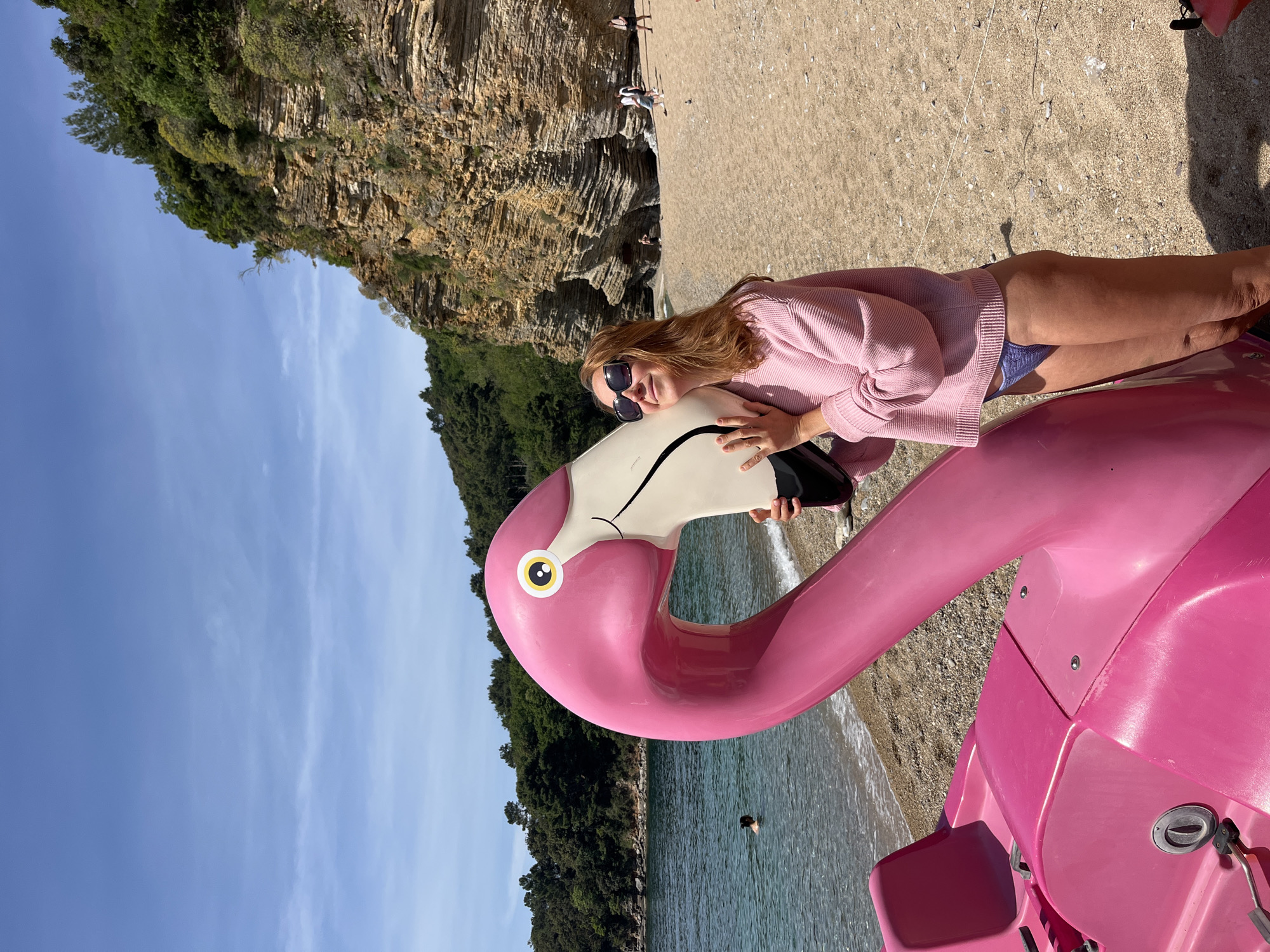 A woman in a pink sweater and dark sunglasses is on the beach in Budva, Montenegro, leaning on a large pink flamingo paddle boat. She is embracing the flamingo's neck. A sandy beach is in the background. Behind the beach, there is a cliff face covered in vegetation on one side and exposed on the other. The sea is visible in the midground, with a person swimming in it. The sky is blue with some wispy clouds.