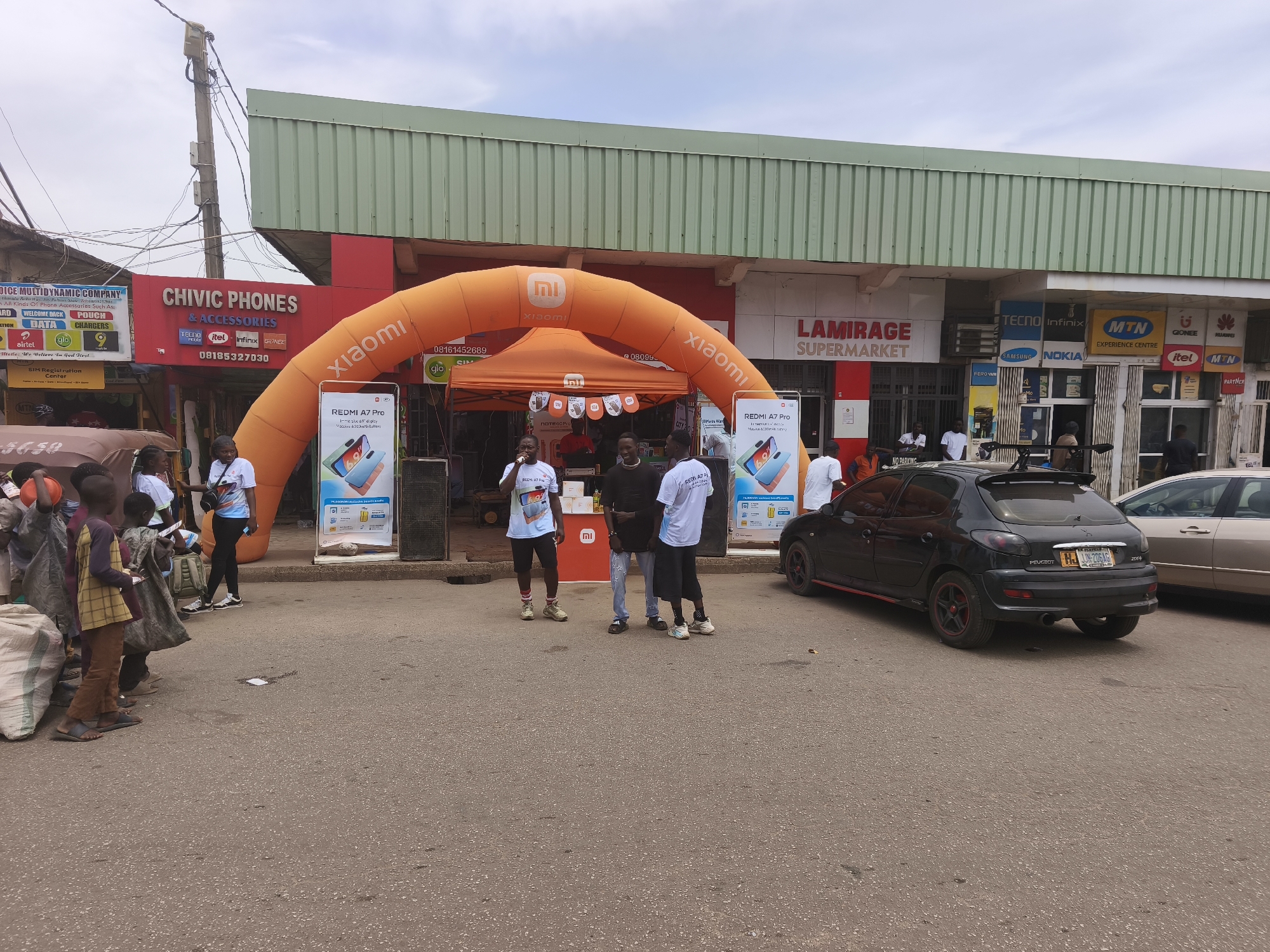 The image captures a street scene in Jos, Nigeria, where a promotional event for Xiaomi mobile phones is taking place. An orange inflatable arch with the Xiaomi logo frames a tent also branded with Xiaomi logos. Two banners displaying the Redmi A7 Pro are positioned on either side of the tent. Several people are present, some wearing T-shirts related to the promotion. A black Peugeot car is parked near the event. Shops with signs like "Chivic Phones" and "Lamirage Supermarket" are visible in the background. Other branded signs for phone networks like MTN, Tecno, and Nokia are also visible, indicating this is a commercial area likely related to mobile phones and services. A group of people appears to be walking down the street. Overhead power lines and utility poles are also visible.