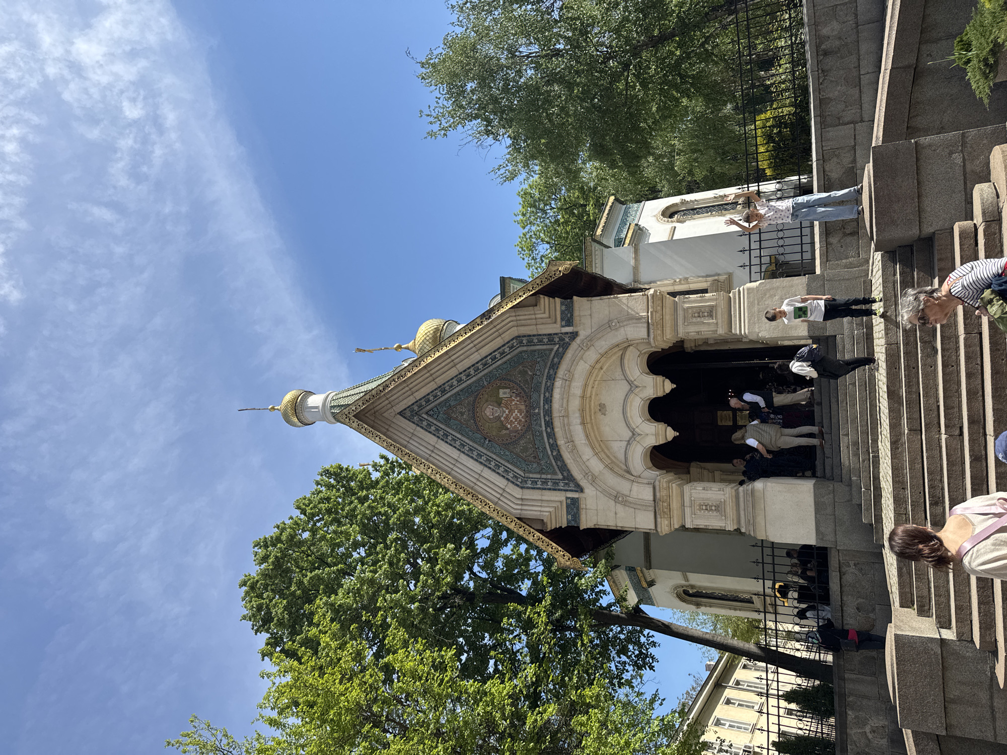 The image shows the Russian Church, a prominent landmark in Sofia, Bulgaria. The church, with its distinctive golden domes and intricate facade, is viewed from a low angle. People are ascending the steps leading to the entrance. The architectural style is Russian Revival, characterized by ornate decorations and vivid colors. Green trees are visible, framing the church against a blue sky.