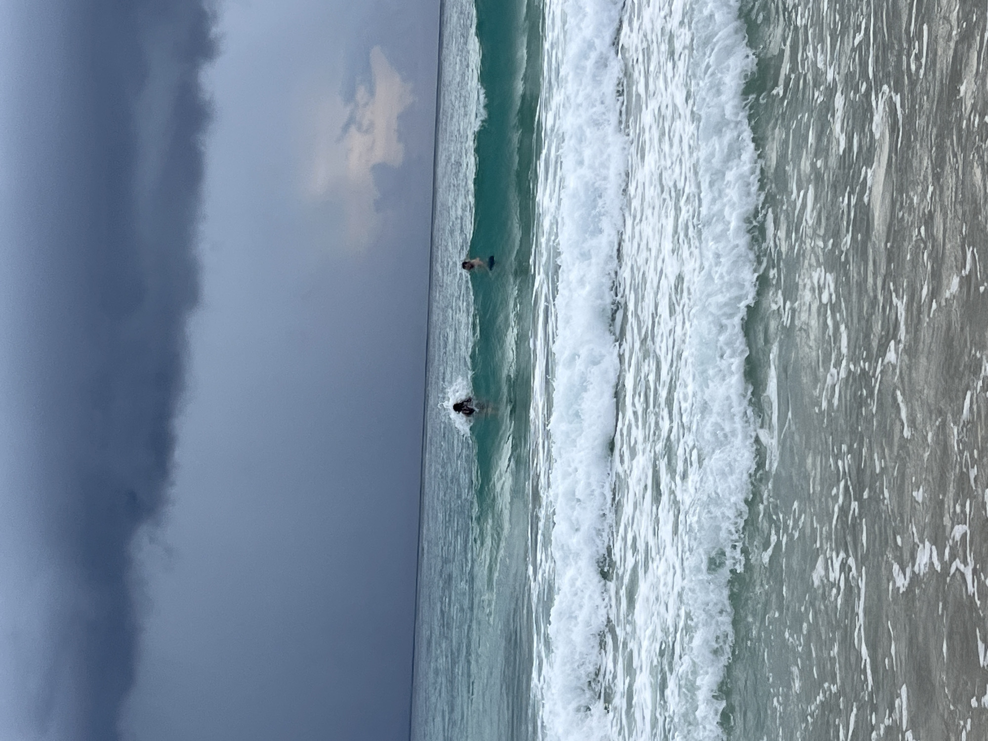 This media file depicts a scenic view of a beach on a cloudy day. The scene is dominated by the ocean with breaking waves and a sandy shore. Two individuals are visible in the water, appearing to be body surfing or engaging in some water sport. One person is closer to the shore, while the other is further out. The sky is heavily overcast with dark clouds, suggesting impending rain or a stormy weather condition. The water is a clear turquoise color, contrasting with the dark sky. The waves are white and foamy, indicating moderate surf. The beach itself appears to be composed of light-colored sand. The image is oriented vertically, with the sky on the left and the ocean on the right. The overall mood of the image is serene, despite the stormy weather, with the focus on the interaction between humans and the ocean. No text is visible in the image.