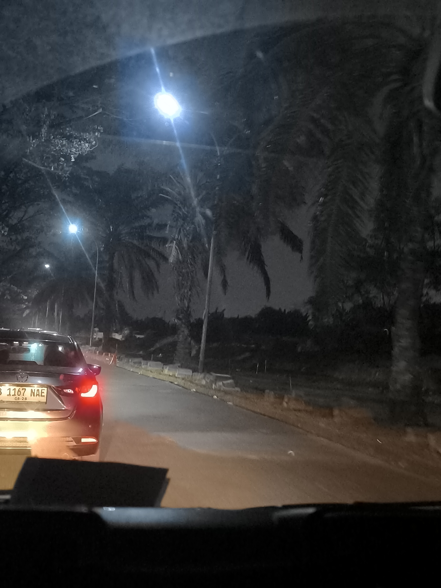 This image captures a nighttime scene from inside a vehicle, looking forward and slightly to the left. The foreground is dominated by the dashboard and windshield of the car, with the view ahead showing a road illuminated by streetlights and the taillights of the car in front. A dark-colored sedan, likely a Mazda based on the emblem on its trunk, is directly ahead, its red taillights illuminated. The license plate of this vehicle reads "B 1167 NAE".
The road appears to be paved and curves to the left. On the right side of the road, there is a raised embankment with some vegetation and palm trees. On the left side, more palm trees and foliage are visible, bordering the road. Several streetlights are present, casting a bright, bluish-white glow. The overall atmosphere is dark, indicative of nighttime. The specific mention of Tangerang, Indonesia, suggests this scene takes place on a street within that city, likely a thoroughfare lined with palm trees and experiencing typical urban nighttime lighting. There are no people clearly visible within the frame.