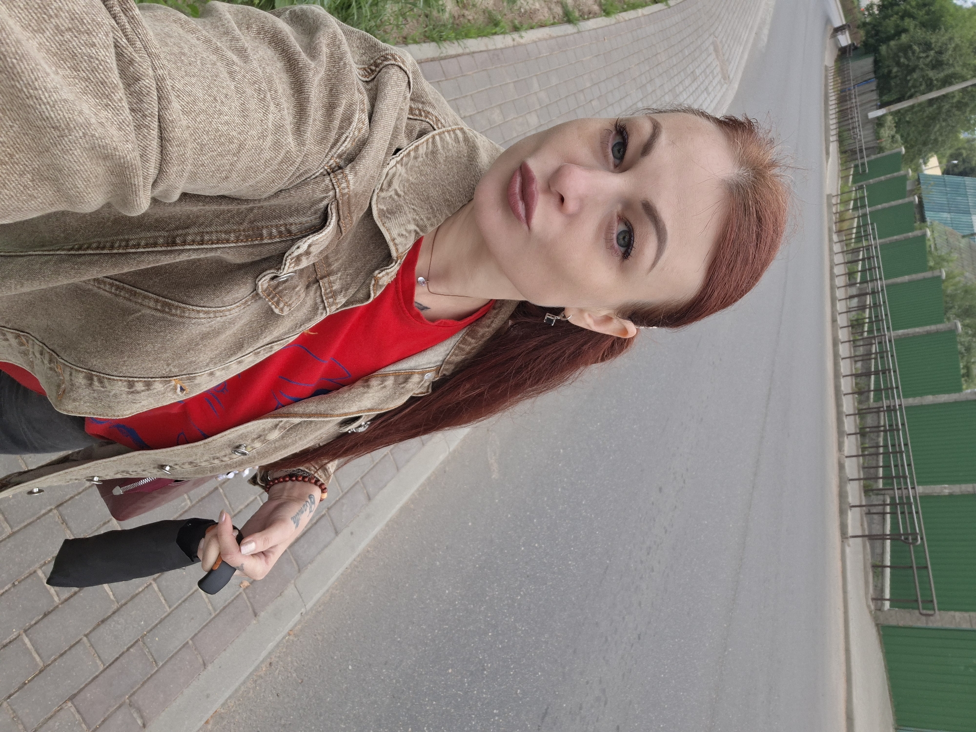 A young Caucasian woman with long, reddish-brown hair and blue eyes is standing on a sidewalk next to a road, holding a folded black umbrella. She is wearing a tan denim jacket over a red t-shirt. The sidewalk is made of interlocking pavers, and there is a grassy area to the left. The road is paved and appears to be a street in a town, with a green metal fence and buildings visible in the background. The scene is outdoors and appears to be daytime. The woman is looking at the camera and has her lips puckered. She has tattoos on her left arm. The location is indicated as Smolya Vichi, Belarus.