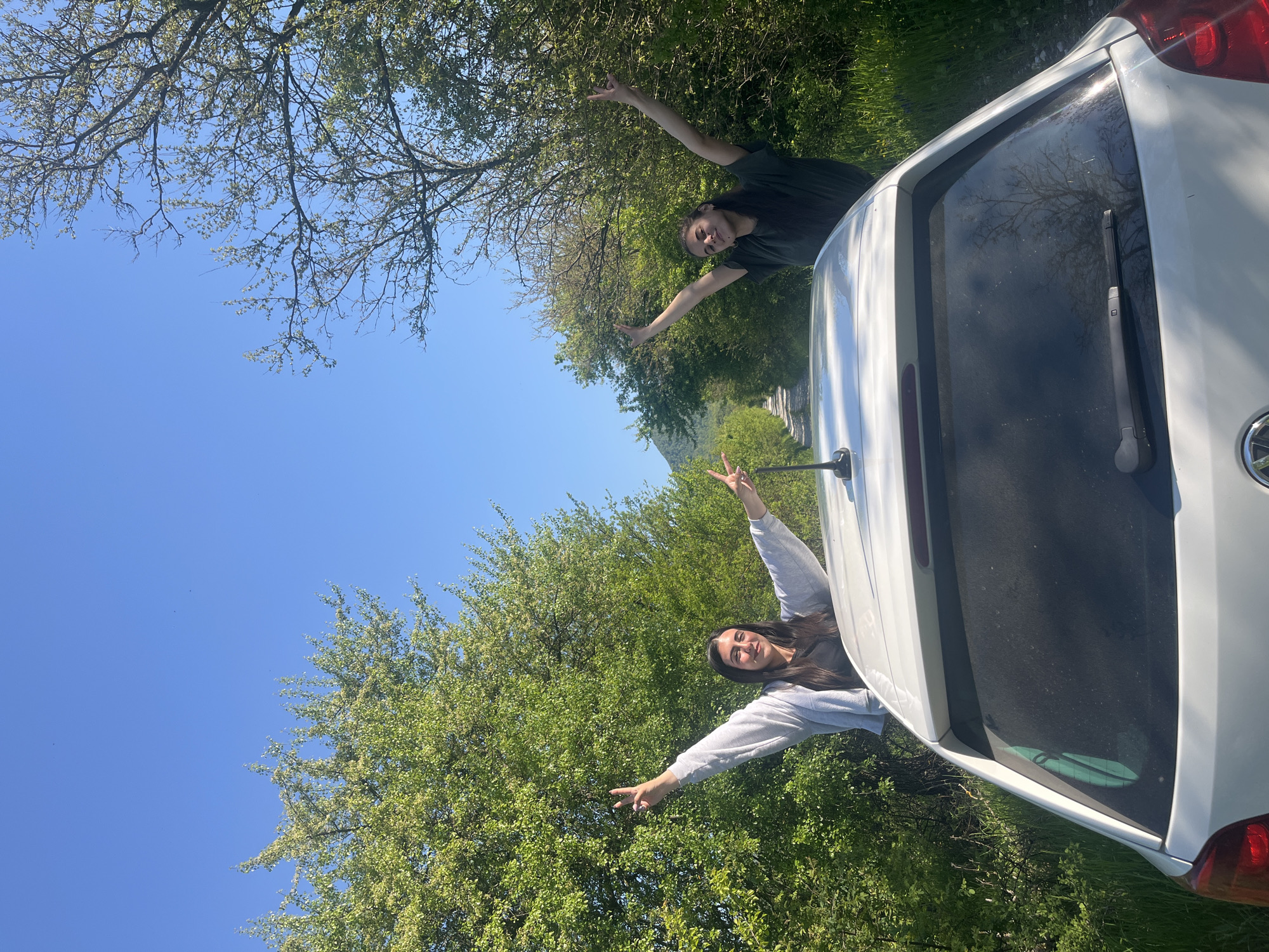 Two women stand beside a white car on a grassy area with trees and a clear blue sky above. Both women extend their arms and make peace signs with their fingers. The location is Rish, Bulgaria, as inferred from the image context.