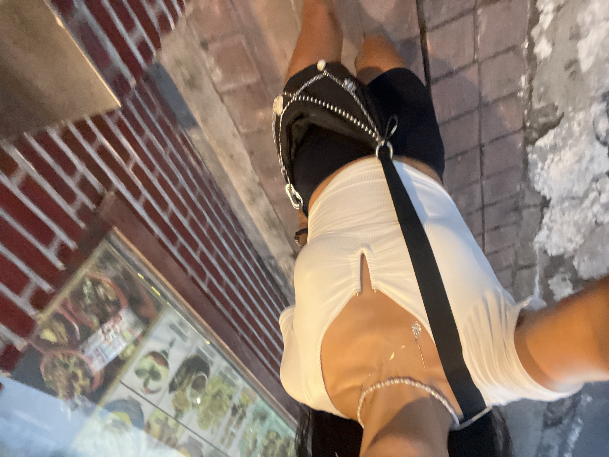 A person stands on a stone-tiled sidewalk. The person wears black shorts and a white, form-fitting top. A black purse or bag is draped across their body. A jewelry store front is to the left, with a glass display case exhibiting various items inside. The person's body is angled and slightly turned to the right. A portion of a brick wall is visible above the store window. The location cannot be confirmed as Kuta Selatan, Indonesia, with only the provided image.