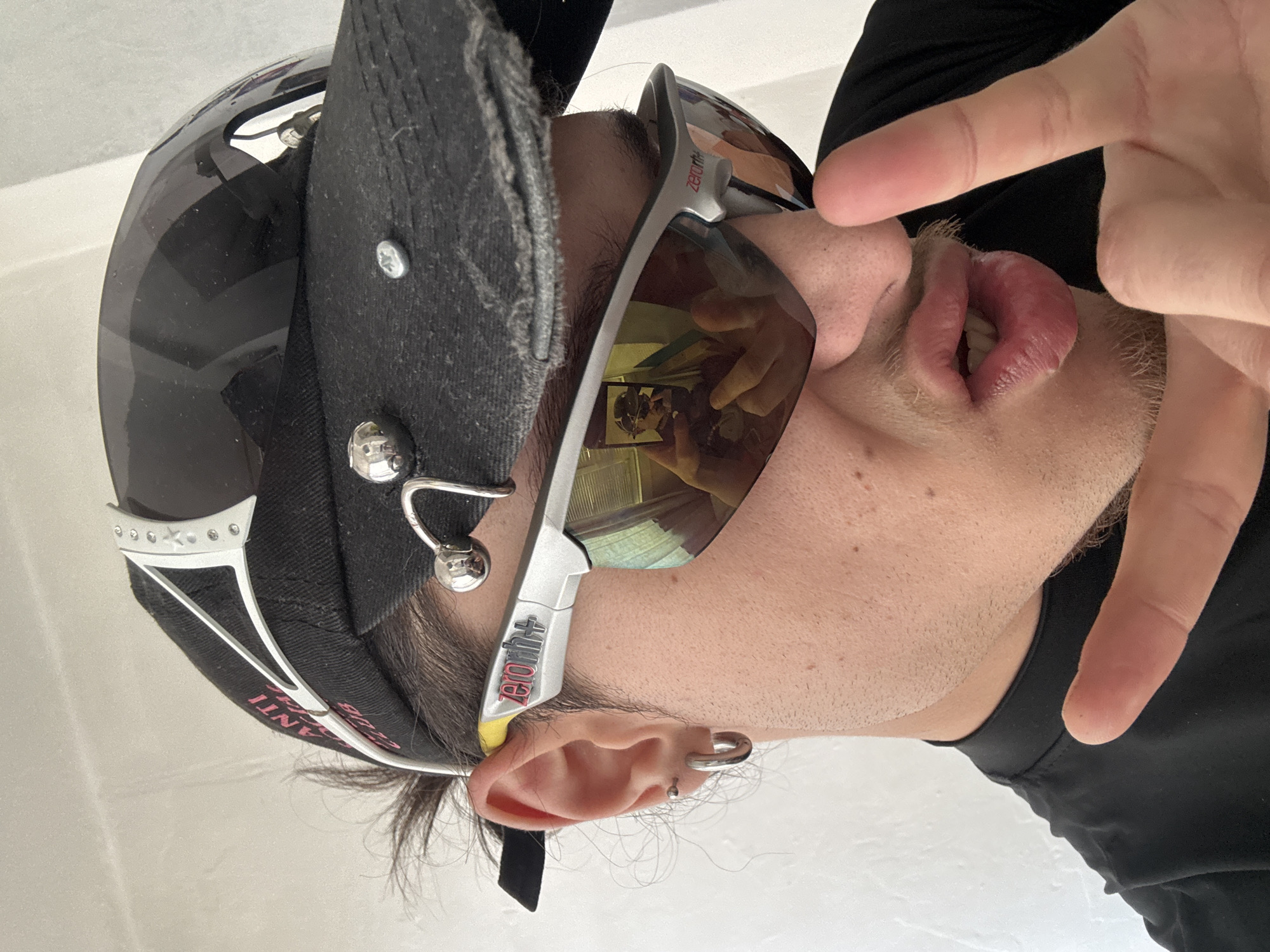 A person is pictured from a low, upward angle wearing sunglasses with reflective lenses, a dark-colored baseball cap with a silver safety pin and metal ball attachments, and a black shirt. They have visible ear piercings and are making a gesture with their fingers near their face. The reflection in the sunglasses shows the person holding a phone, suggesting a selfie. The background is a plain white surface. There is no information present in the image that allows to determine the specific location in Sofia, Bulgaria.