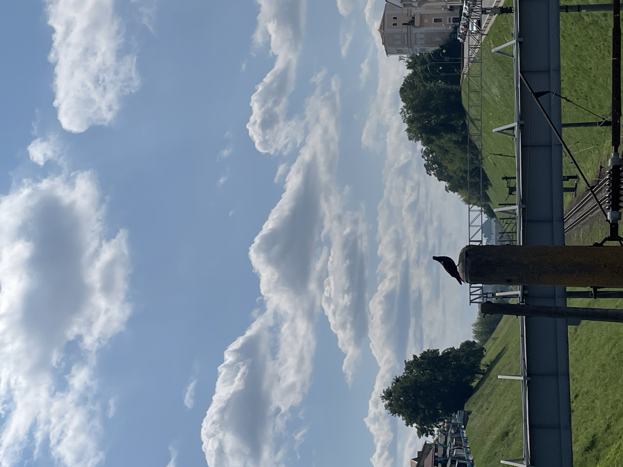 A pigeon is perched on a concrete post, silhouetted against a blue sky with scattered white clouds. The post is in the foreground, and behind it, a metal bridge spans across a railway track. The scene appears to be in an urban environment, possibly Hrodna, Belarus, with grassy slopes and trees lining the sides of the railway. A building with arched windows and a red-tiled roof is visible in the background to the right, along with parked cars on a road. The lighting suggests it is daytime.