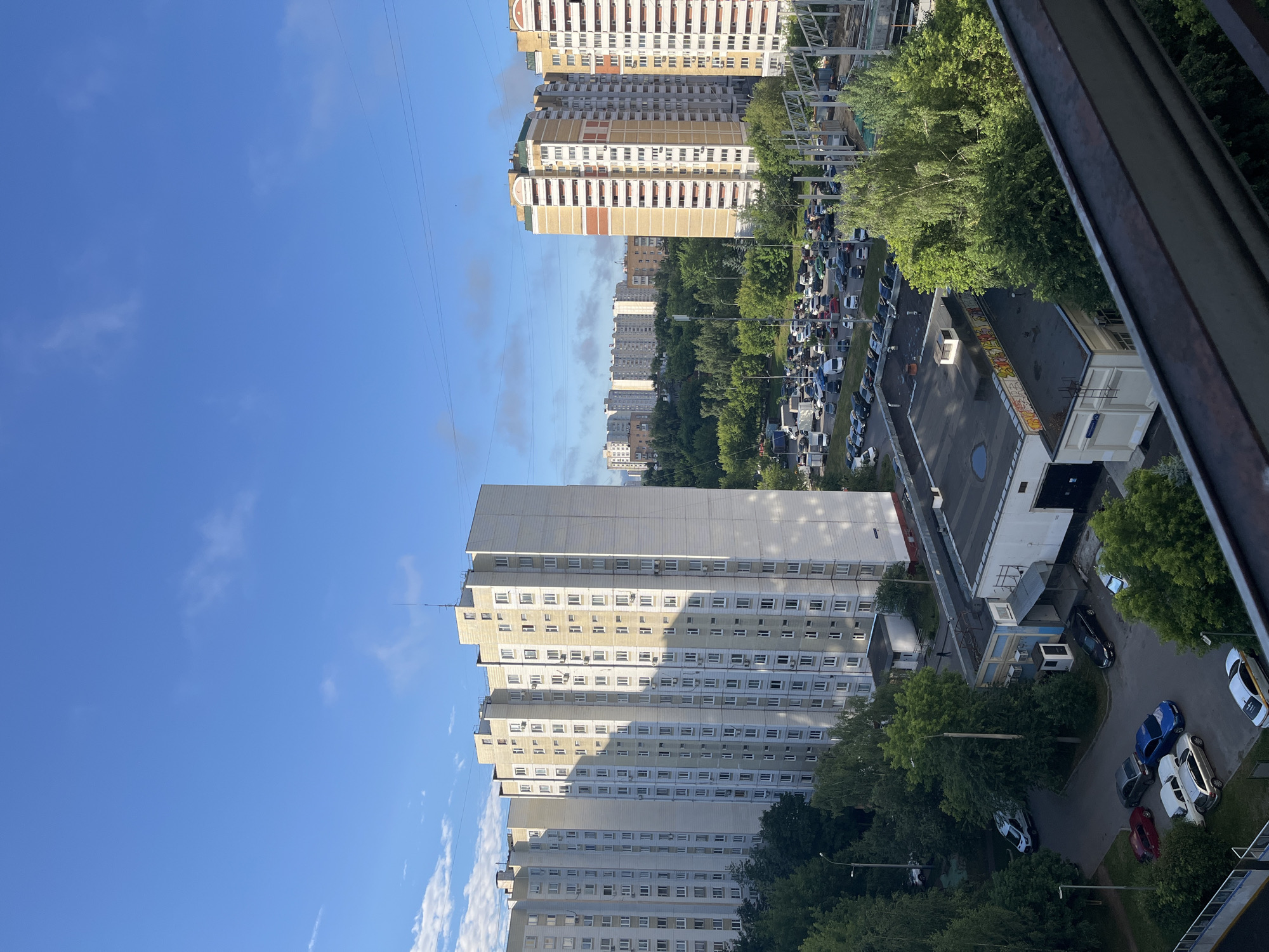 This image depicts a city scene in Moscow, Russia, likely during the daytime. The foreground is dominated by a dark, weathered railing, suggesting the vantage point is from a balcony or elevated structure.
The midground reveals a cluster of Soviet-era panel buildings, characterized by their repetitive modular design and light-colored facades. These buildings are tall and densely packed, typical of residential areas in Moscow. Interspersed with the buildings are mature trees, casting shadows on the facades and indicating a green urban environment.
In front of the panel buildings, a busy street is visible, lined with parked cars of various makes and models. The sheer number of vehicles suggests a residential or mixed-use area with ample parking. Beyond the residential blocks, more buildings are visible in the distance, extending the urban sprawl.
A construction site is also present, with scaffolding and incomplete structures hinting at ongoing development or renovation. The sky is blue with scattered clouds, and power lines are visible crisscrossing the frame, a common element in urban landscapes. The overall impression is one of a functional, lived-in urban environment, typical of many Moscow neighborhoods.