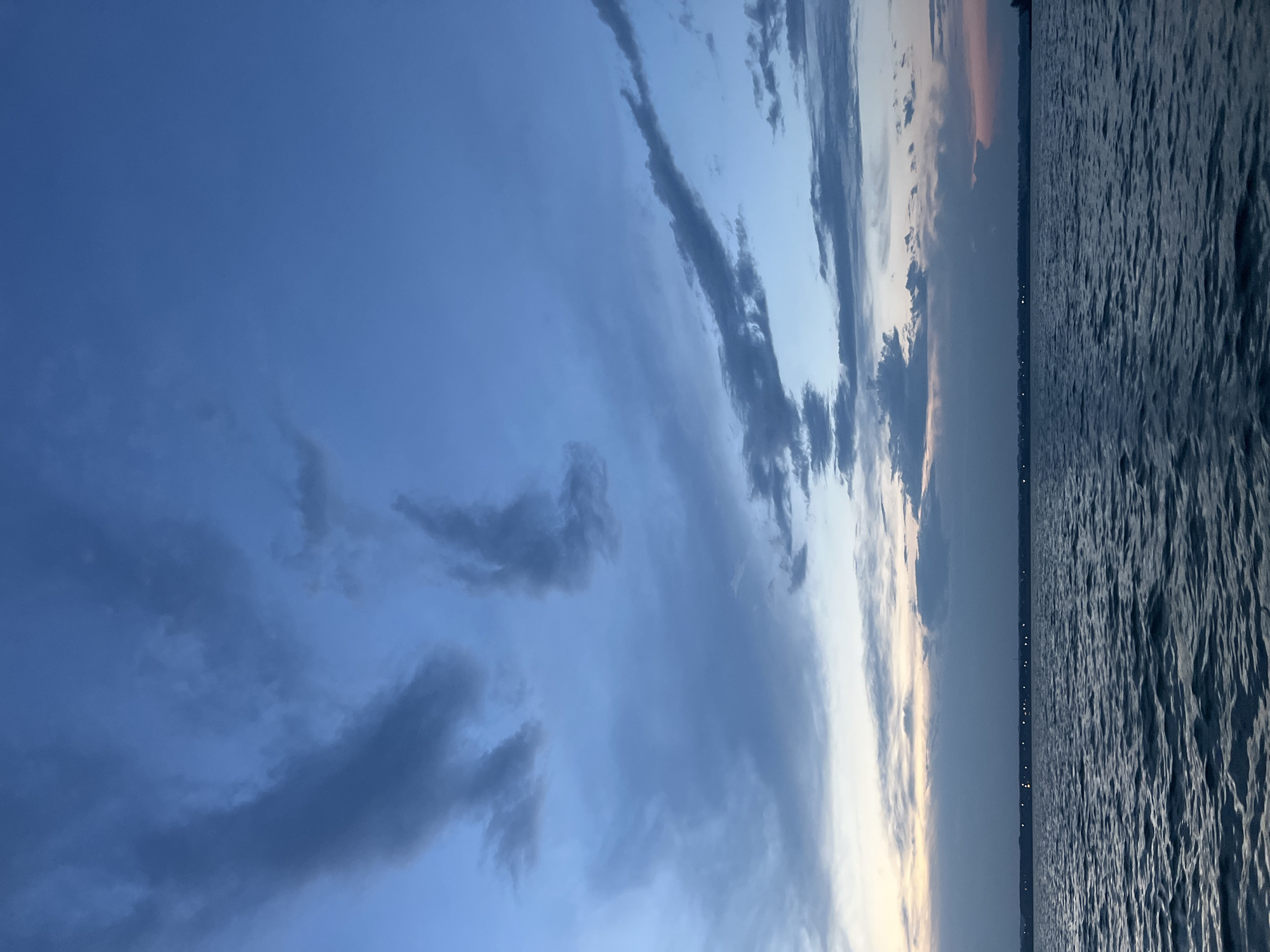 The image captures a serene twilight scene over a body of water, likely a lake or a wide river, with a distant shoreline. The sky is a deep, moody blue, transitioning to lighter shades near the horizon where faint streaks of pale orange and pink suggest the last vestiges of sunset. Scattered, dark grey clouds drift across the upper expanse, some appearing wispy and elongated, others more dense and textured.
The water's surface is rippled, reflecting the muted sky tones. A thin line of lights, possibly from a town or settlement, is visible along the distant shoreline, indicating human presence. The overall atmosphere is calm and tranquil, with a sense of impending nightfall. There are no people or explicit actions depicted in the scene.
