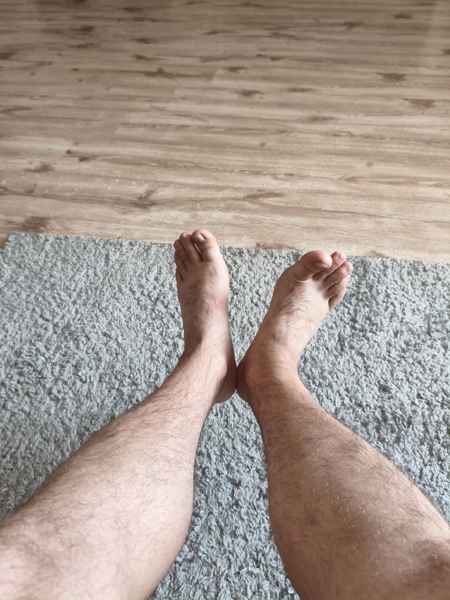 The image shows a top-down view of two bare, hairy legs and feet resting on a grey textured rug. The rug is placed on a wooden floor with planks arranged horizontally. The lighting suggests an indoor environment. The image does not provide any information about the city of Sibay, Russia.