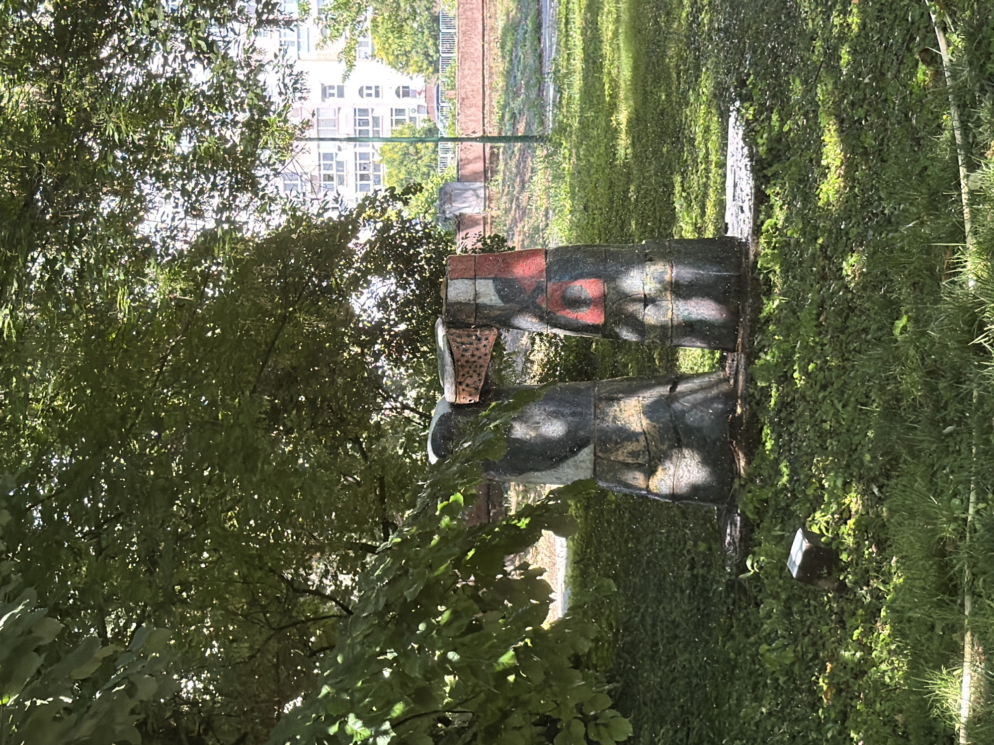 The image shows a weathered, abstract sculpture in a park-like setting. The sculpture appears to be made of rough-hewn stone pillars, painted with abstract shapes and colors, including red and black. These pillars are connected at the top by a rusty metal element. The sculpture is situated on a concrete base in a grassy area with lush green vegetation. In the background, a large tree with dense foliage casts dappled shadows on the sculpture and ground. Beyond the trees, a multi-story building with numerous windows is visible, suggesting an urban environment. A low brick wall and some fencing are also present in the background, and a light pole stands near the building. The lighting suggests it is daytime, with sunlight filtering through the leaves. This sculpture is located in Pazardzhik, Bulgaria, specifically in the park surrounding the Regional Historical Museum.