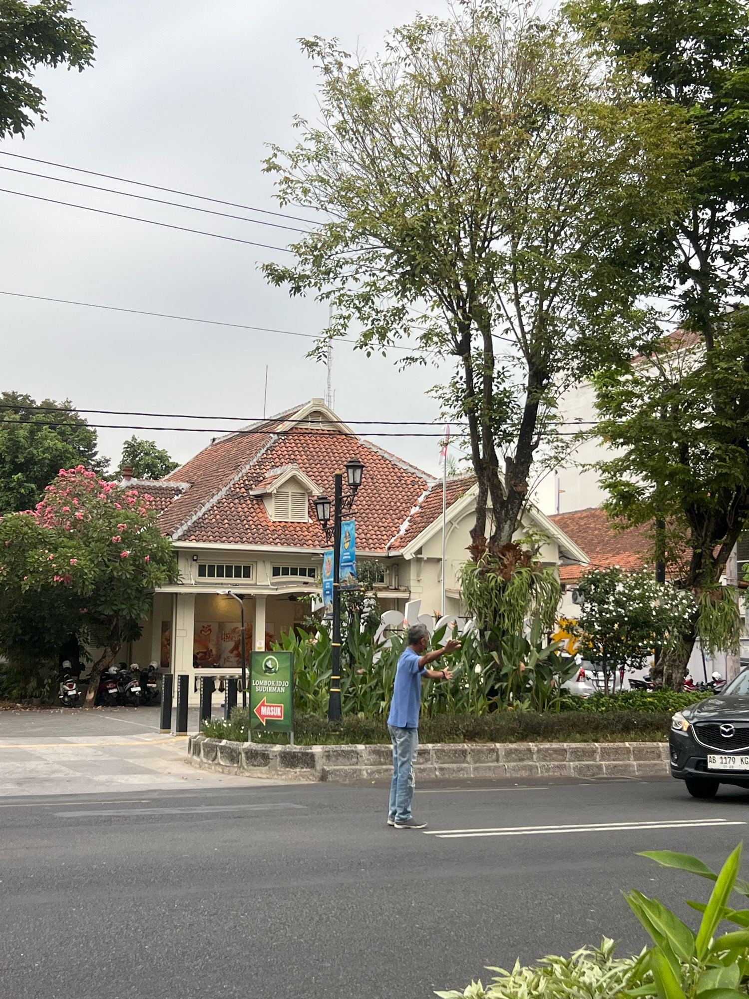 The image captures a street scene in Yogyakarta, Indonesia, on an overcast day. In the foreground, a man in a blue t-shirt and ripped jeans stands in the middle of the road, with his arms outstretched, appearing to direct traffic or engage with a vehicle. A dark-colored car, partially visible on the right side of the frame, has a license plate that reads "AB 1179 KG". Behind the man and the road, a building with a terracotta tiled roof and white walls is visible, featuring arched windows and dormer windows. The building is surrounded by lush greenery, including trees with pink blossoms and various plants. A signpost with green and red signage is located near the entrance of the building, with text including "LOMBOK IDJO SUDIRMAN" and "MASUK" with an arrow pointing left. Several motorcycles are parked to the left of the building. Power lines are strung across the sky, and tall trees with dense foliage frame the upper portion of the image.