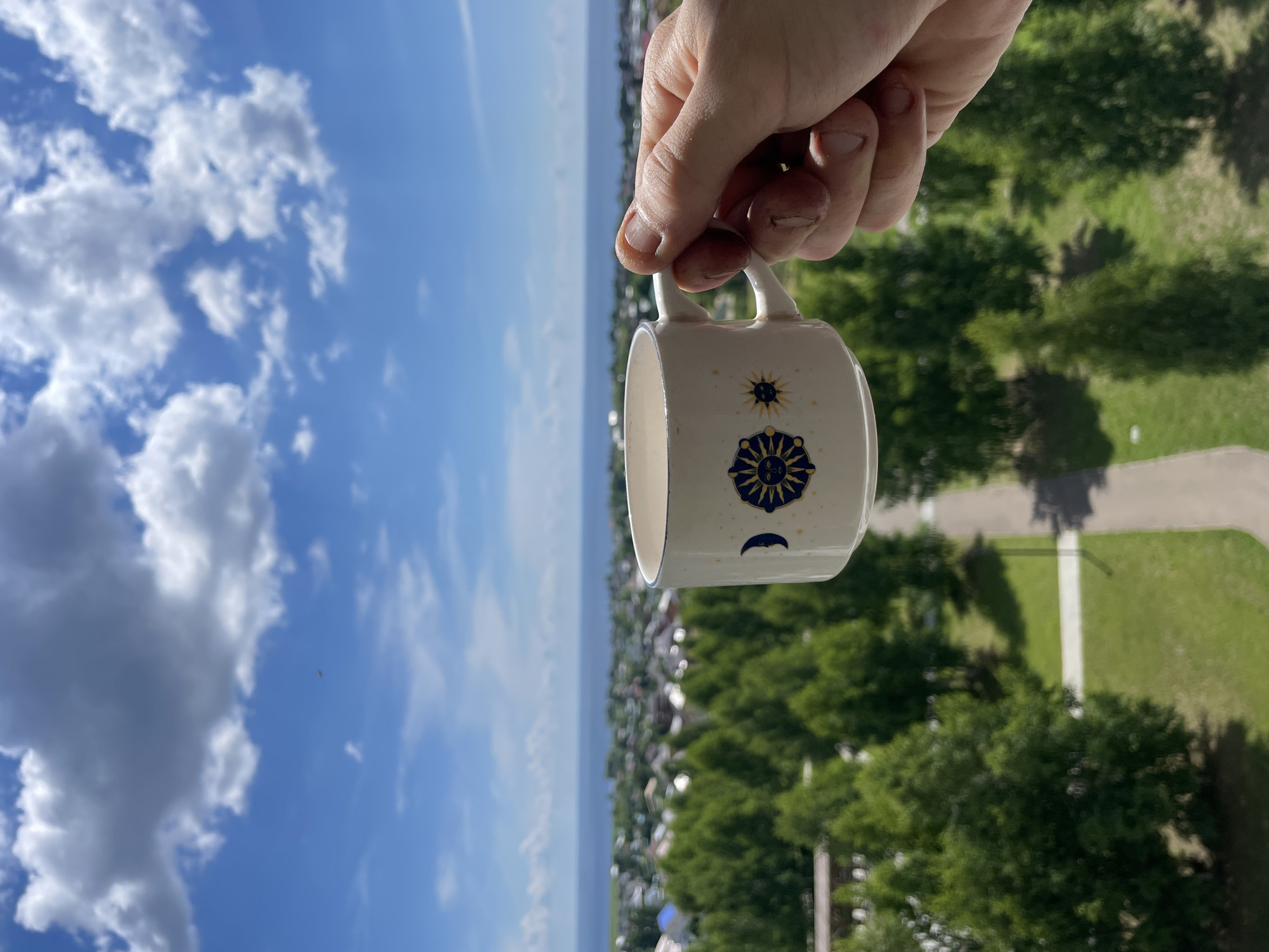 A hand holds a white mug decorated with celestial symbols in the foreground. The mug is positioned against a backdrop of a blue sky with scattered white clouds, a body of water, and a cityscape with green trees. The scene appears to be from a height, overlooking the city of Laishevo, Russia. The focus is sharpest on the mug and the hand holding it, with the background gradually softening into a blur. The lighting suggests a bright, sunny day.