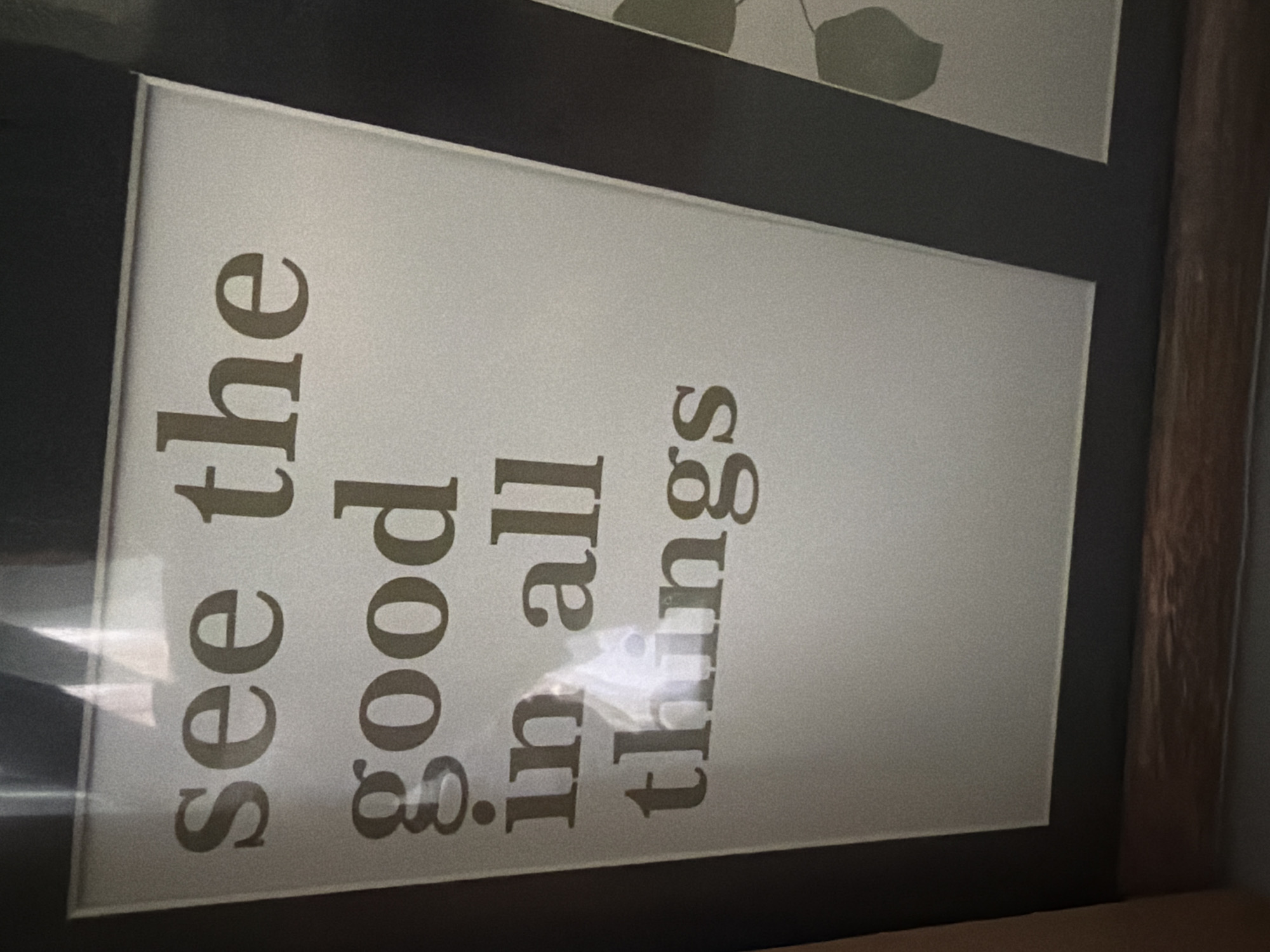 The image shows a framed motivational quote in Bulgarian. The quote reads "see the good in all things". The text is printed in a serif font on a light-colored background, which appears to be a matte paper or cardstock. The frame is dark, possibly black or dark brown, and there is a wooden element visible at the bottom of the frame. The image is slightly blurry and tilted, with some reflections on the surface of the frame, suggesting it is a photograph of a framed item. The background outside the frame is dark and out of focus. There is no indication of the location being Pazardzhik, Bulgaria, or any other specific city.