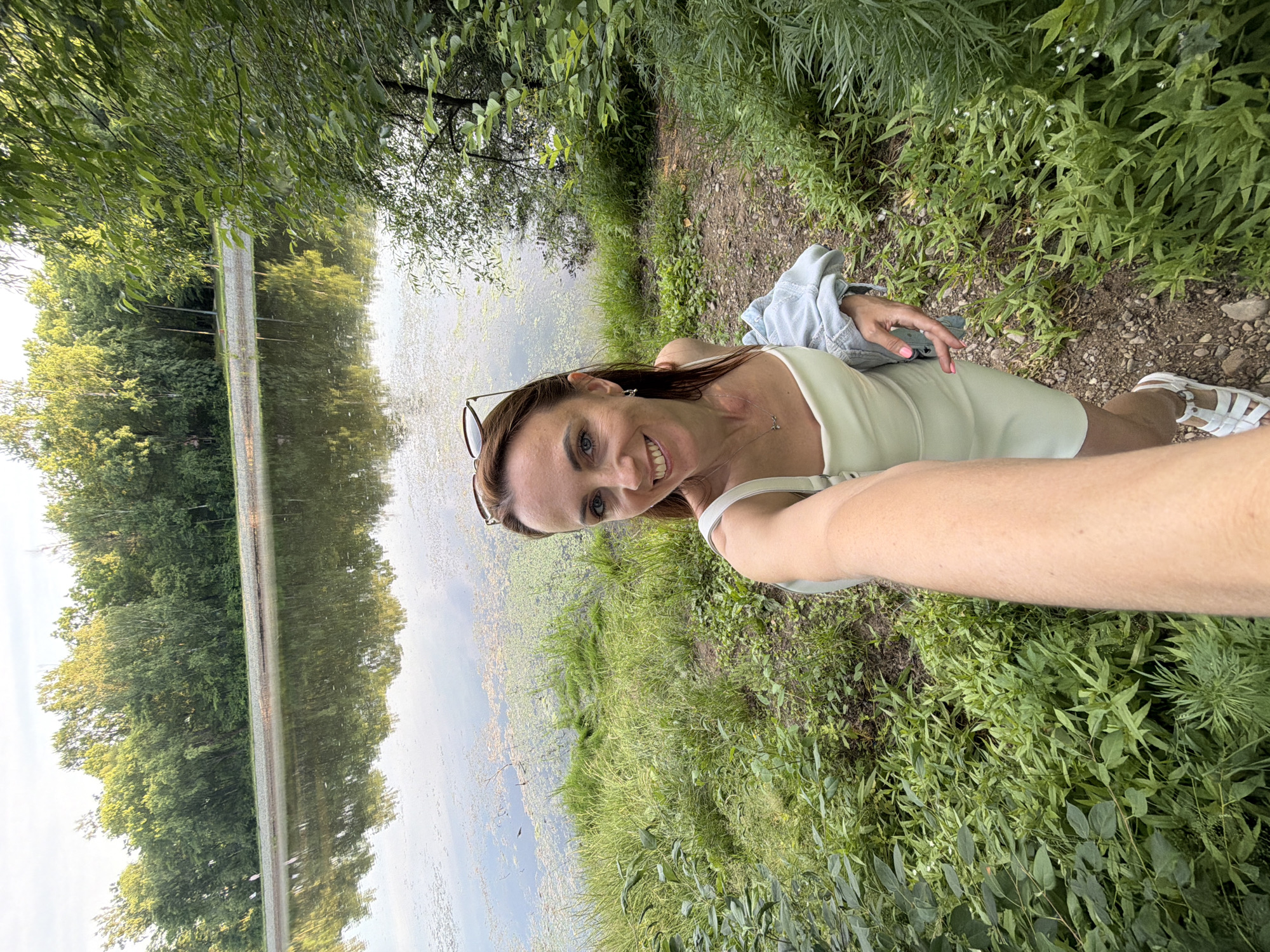 A woman takes a selfie while standing on a dirt path next to a body of water. She is wearing a light green tank top, a light blue jacket, and white sandals. She has sunglasses perched on her head and is smiling at the camera. The body of water is calm and reflects the trees and sky. There is dense green foliage on the right side of the path and the water. The overall scene suggests a peaceful outdoor setting. The time of day appears to be daytime, with bright natural light. The woman's expression conveys happiness and a sense of enjoyment.