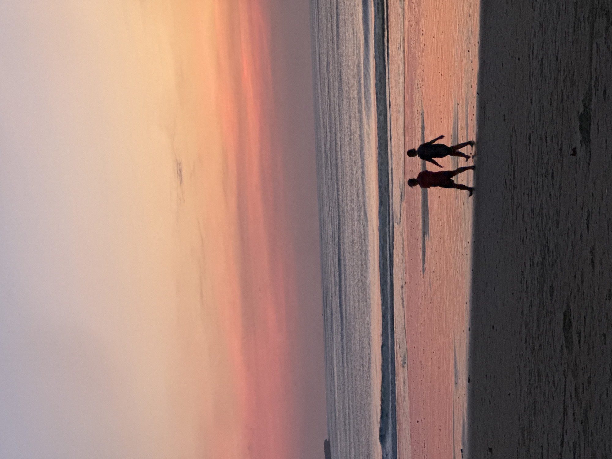 A silhouette of two individuals walking on a beach during sunset is captured in this photograph. The sky displays a gradient of colors, transitioning from pale lavender at the top to soft pinks and oranges near the horizon. The ocean stretches out, with gentle waves lapping at the shore, reflecting the warm hues of the sky. The wet sand on the beach also mirrors the sunset colors. The foreground is dominated by dry sand, casting a shadow from the setting sun. One person appears to be wearing shorts and a t-shirt, while the other is in athletic wear. The location is Kuta Selatan, Indonesia.