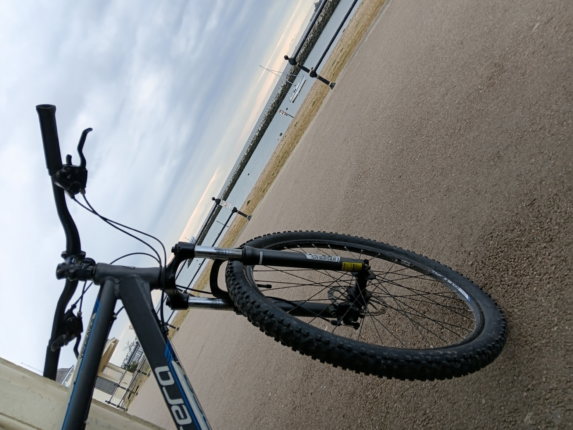 The image shows a black and blue Carrera Vengeance mountain bike from a low, angled perspective, focusing on the front wheel and handlebars. The bike is on a paved surface, likely a road or path. Beyond the path is a body of water, possibly a bay or harbor, with a breakwater visible in the distance and a few boats. The sky is overcast. Based on the breakwater structure and presence of boats, and given the city context, it is likely that the location is near the coast of Canterbury, United Kingdom. The bike is stationary in the foreground. There are no people visible in the image.