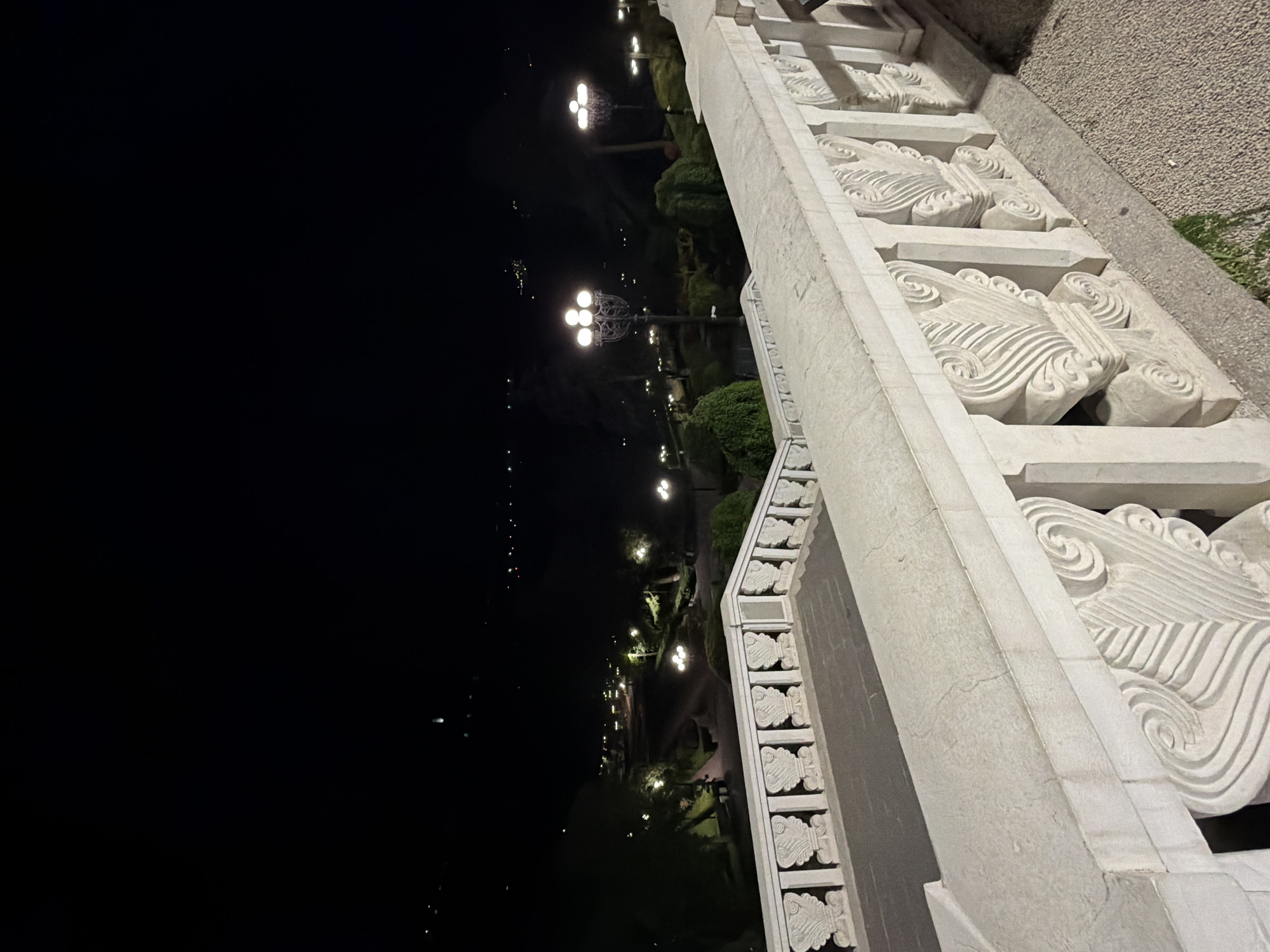 The image shows a section of a carved white stone balustrade at night in Tbilisi, Georgia. The balustrade is highly ornamented with a repeating, curvilinear design. Adjacent to the balustrade is a gravel or paved surface with some green vegetation visible. In the background, a row of illuminated street lamps lines a pathway, suggesting a park or promenade. Further in the distance, there are sparse lights, hinting at the presence of a city skyline or other urban elements. The foreground is mostly black, indicating night conditions and a limited light source. The image captures a static scene with no clear people or actions directly visible.