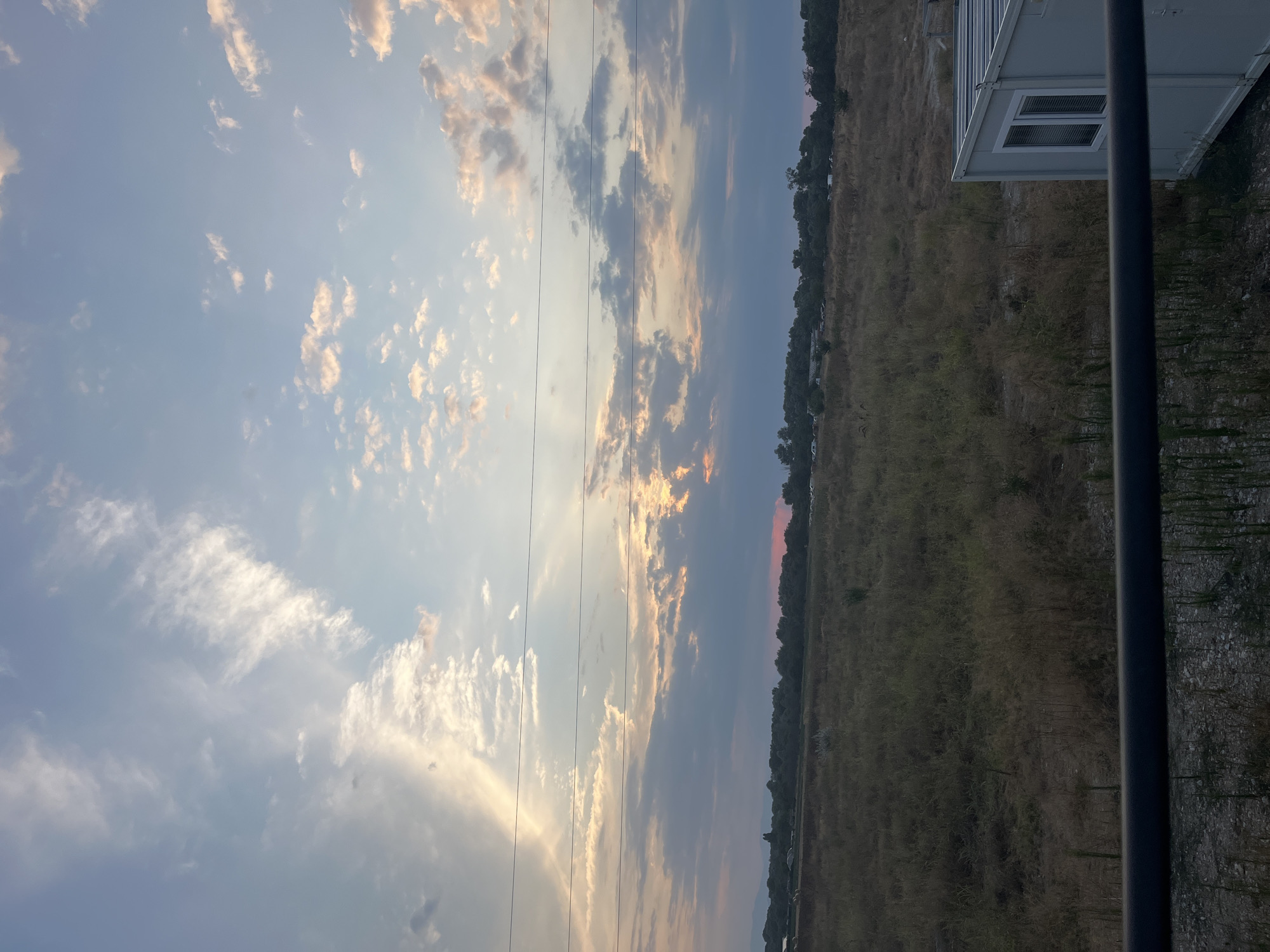 The image captures a sunset over a rural or semi-rural area, possibly on the outskirts of Plovdiv, Bulgaria. The sky is filled with dramatic clouds, tinged with the warm colors of the setting sun. Rays of light pierce through a gap in the clouds, illuminating the sky with a soft glow. In the foreground, a field of dry, tall grass stretches towards a distant treeline. Power lines are visible across the middle of the frame. To the right, a portable cabin or modular building with windows is partially visible. The overall scene suggests a quiet, natural environment at dusk.