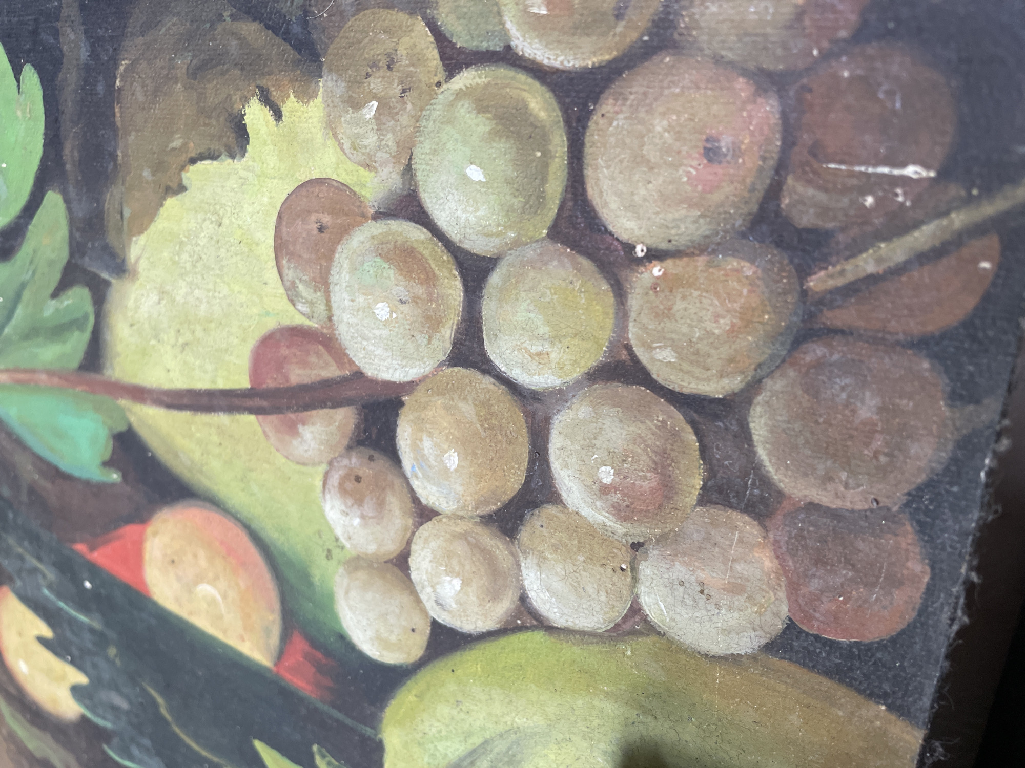 This is a close-up, eye-level, indoor shot of a painting depicting a still life of fruits. The painting features a cluster of grapes in the foreground, with some appearing light green and others a reddish-brown. To the left, a large, elongated green fruit, possibly a pear or apple, is visible. In the background, a hint of a red object, possibly a bird, is peeking through dark green foliage and a pale yellow background. The painting exhibits a textured, somewhat aged appearance with visible brushstrokes and some cracking. The lighting is natural, casting soft shadows on the depicted fruits. The overall impression is one of abundance and rustic charm, typical of still life compositions.